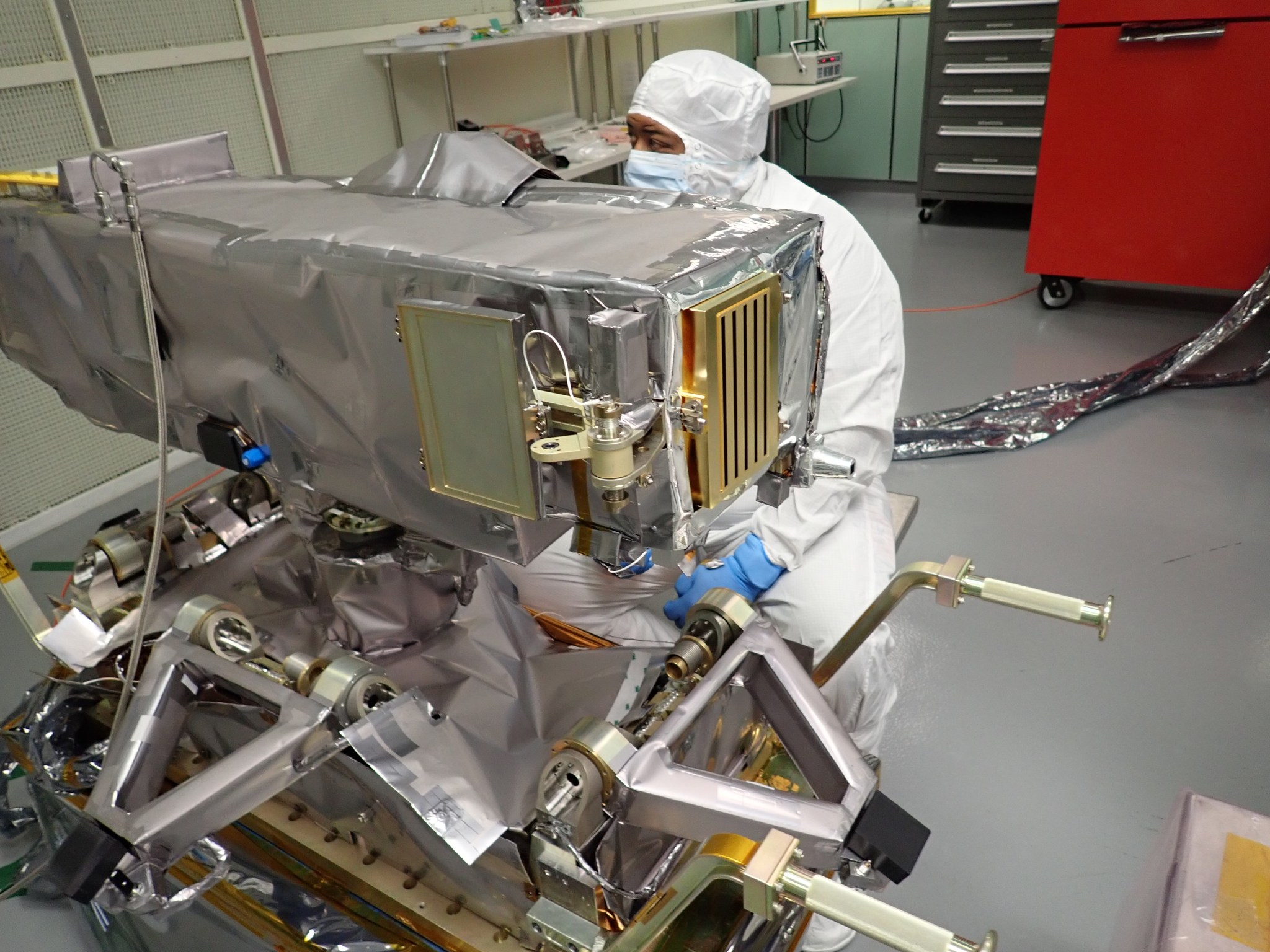 A man wearing clean room gear sits behind a large rectangular instrument, wrapped in shiny silver material. 