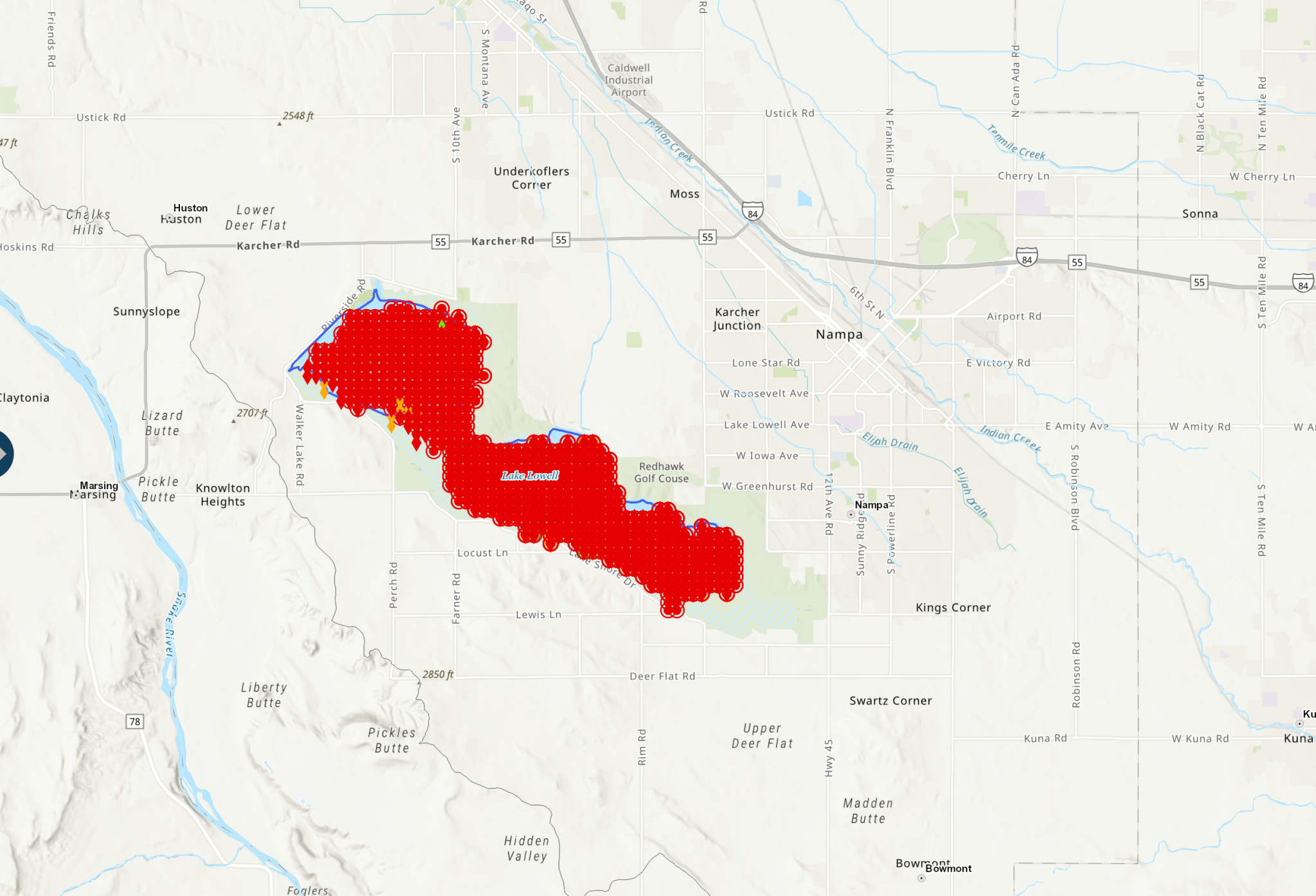 A map of a small lake in Idaho, with bright red dots showing where a cyanobacteria bloom has reached concentration levels that are dangerous to human health. A few small orange and green icons are visible near the lake's edges, indicating areas of lower concentration. The lake is shaped like a thick squiggly line roughly horizontal. The background map is tan and has text and lines showing the nearby towns of Karcher Junction, Nampa, and Knowlton Heights.