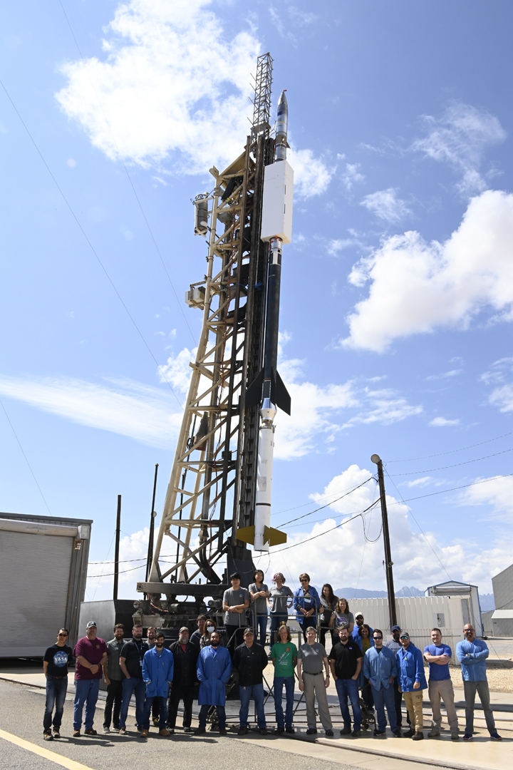 Over two dozen people stand in front of a sounding rocket  mounted on a vertical launch platform.