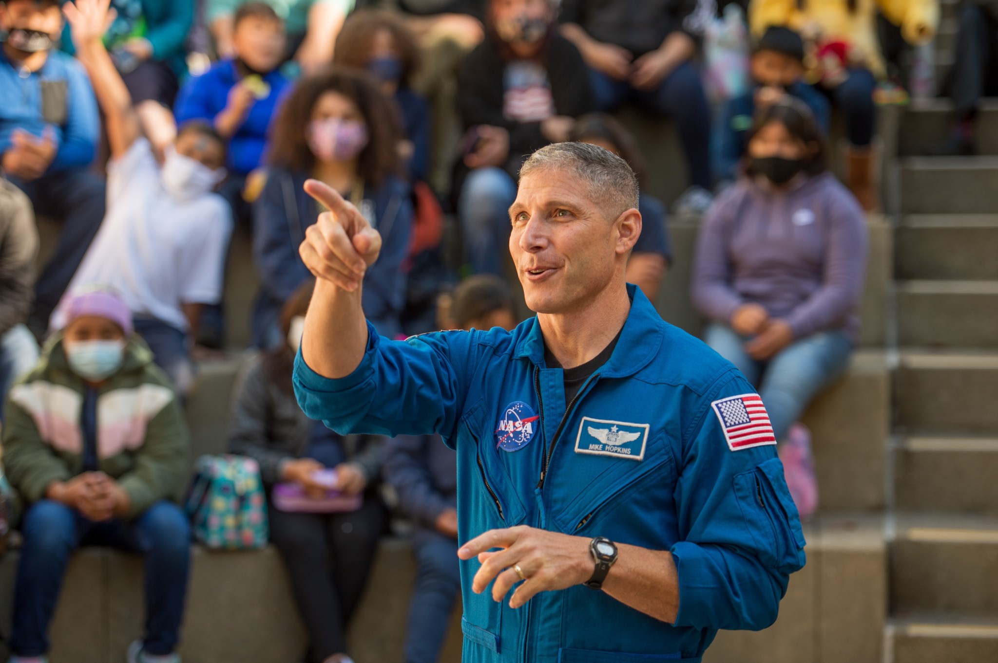 Man in a blue jumpsuit pointing to the left, with an audience of young students blurred in the background.