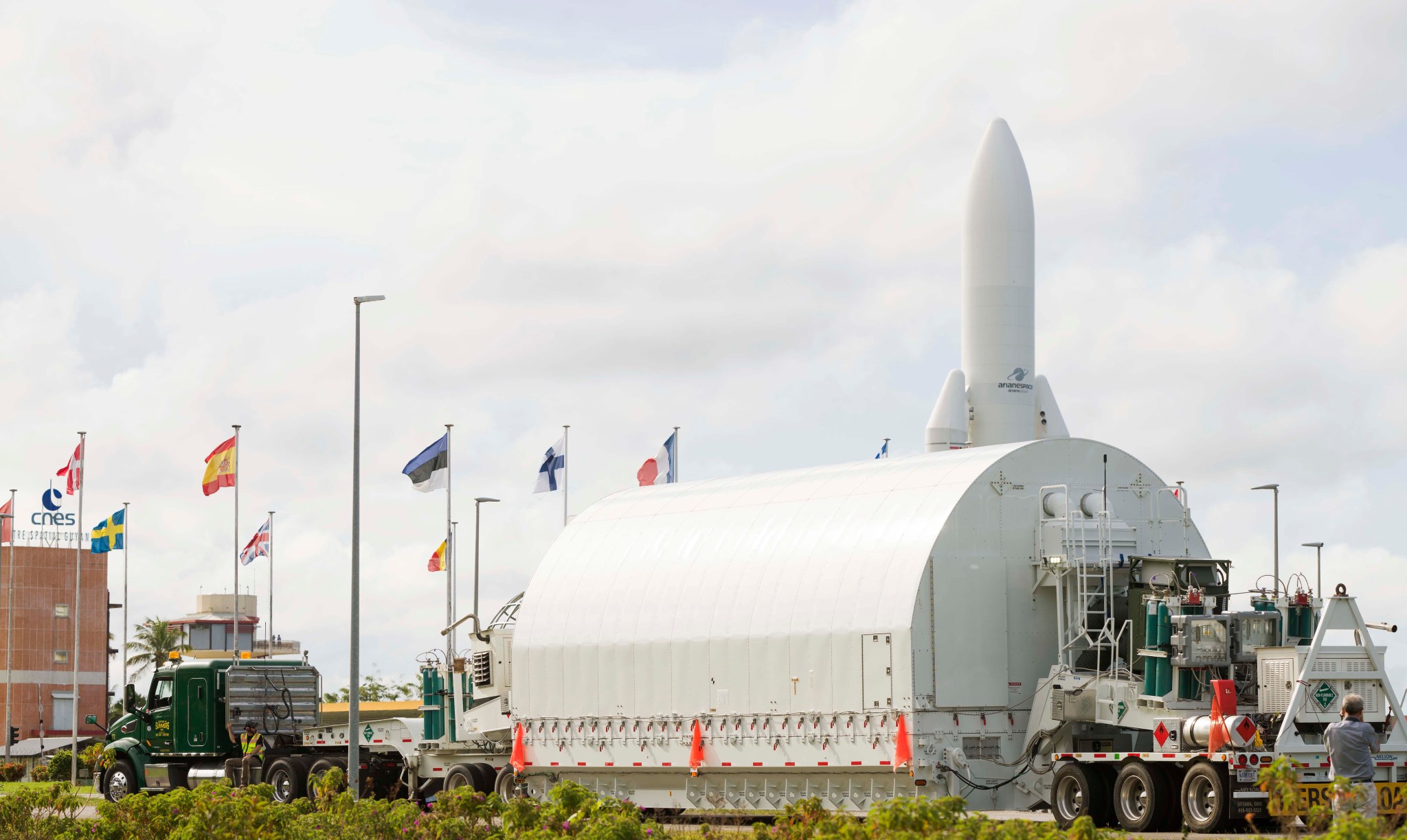 The James Webb Space Telescope inside a white container rounded on top and flat on the bottom on a flatbed trailer. The trailer is passing by a mock white rocket. Multiple countries' flags are also flying along the roadway.