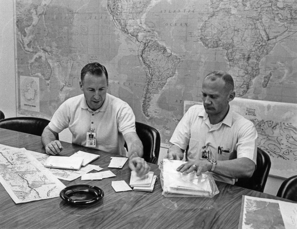 Gemini XII astronauts James A. Lovell, left, and Edwin E. “Buzz” Aldrin sit at a table to review flight documents. A map of the world appears behind them.
