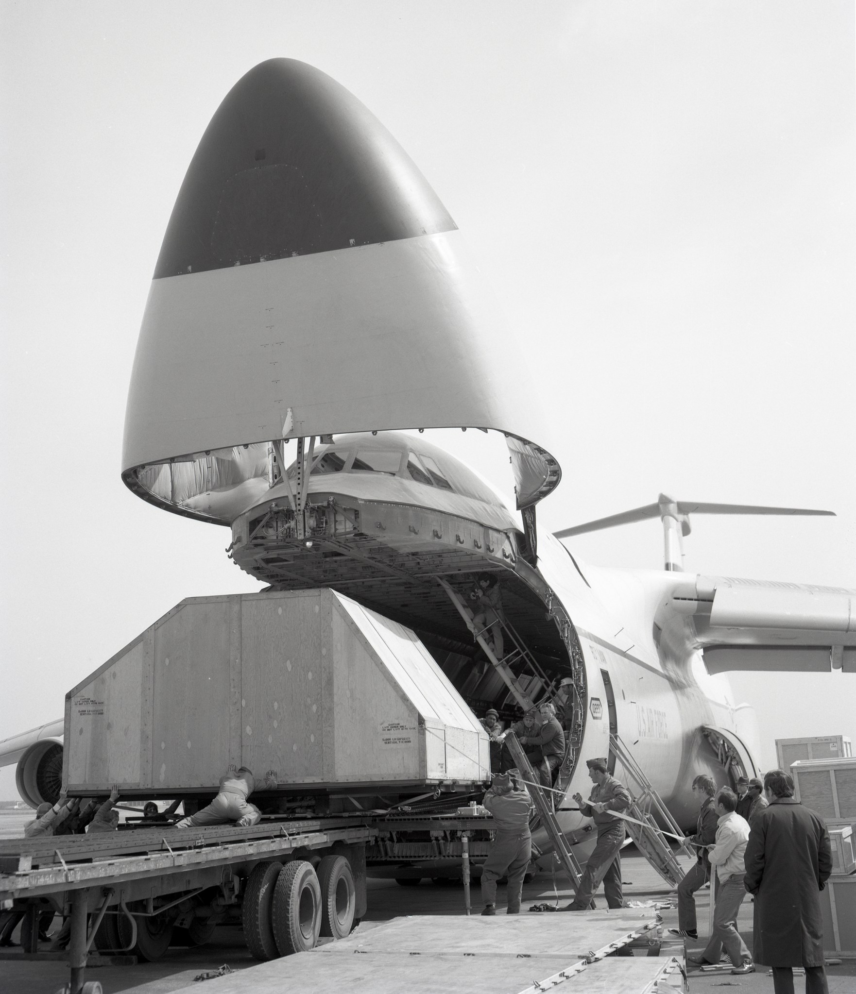 Hinged nose of large airplane is tilted up revealing a large wooden crate. Workers poised to remove to a flatbed truck.