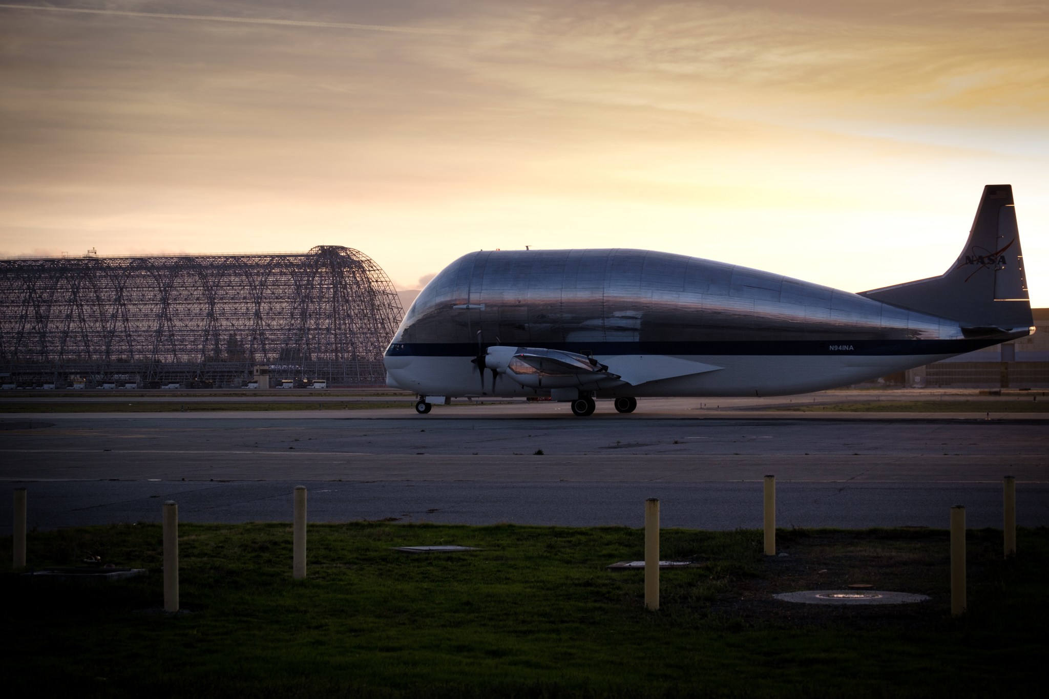 Plane taxiing on an airfield, with the structure of a decommissioned hanger in the background. Sun is rising in the background.