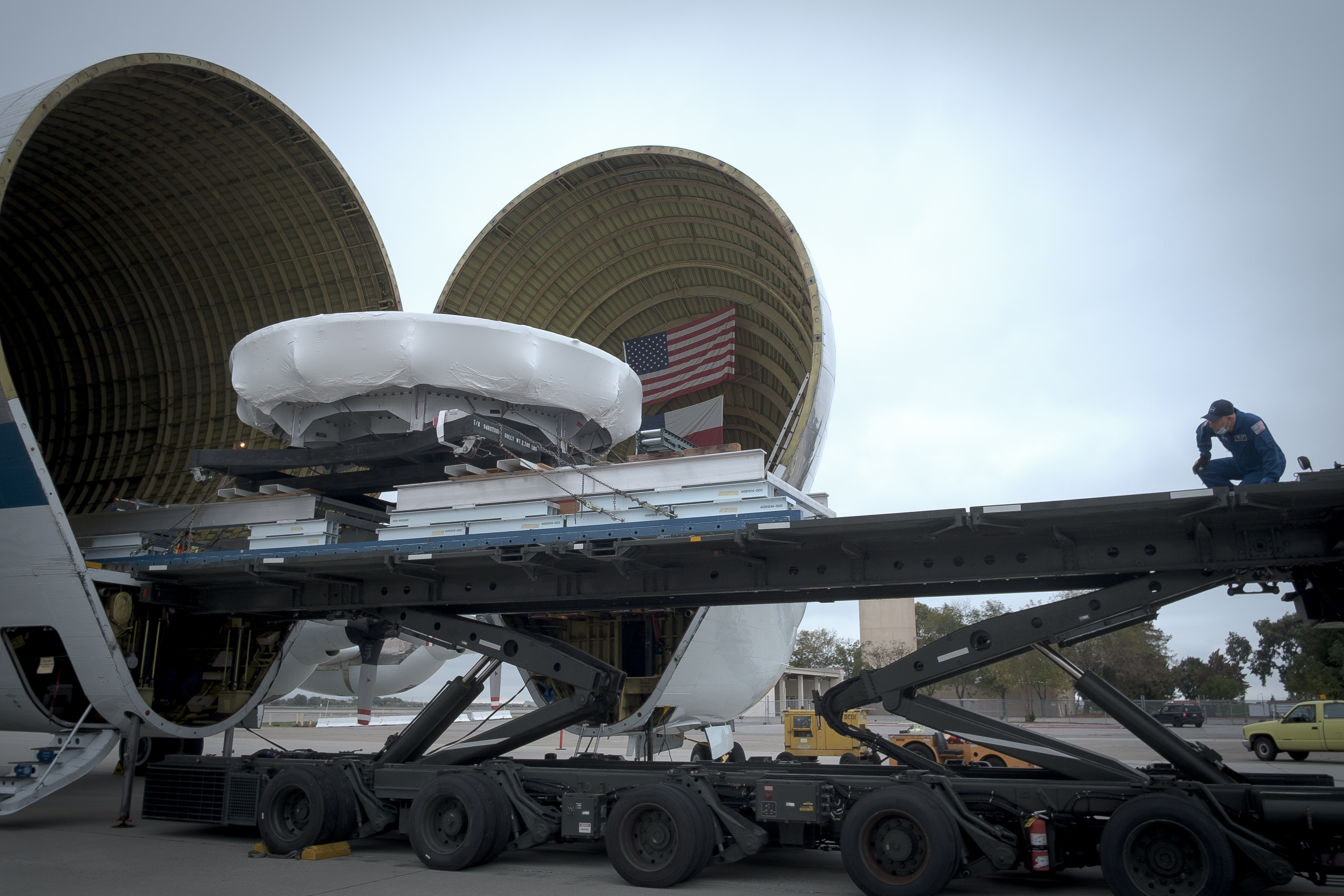 A large plane is open, with the front nose split, and the circular heat shield skin being rolled out from inside. The sky is grey.