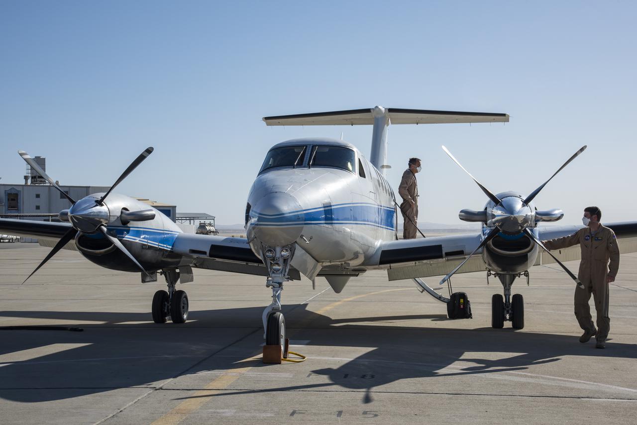 Two members of a flight crew wear brown coveralls and inspect a small silver plane with a blue stripe down its center. One of the crew members stands on the plane's wing, and the other stands at the far right of the image with their hand on the plane's engine. The small plane has two engines and sits on a runway, with a building visible in the background.