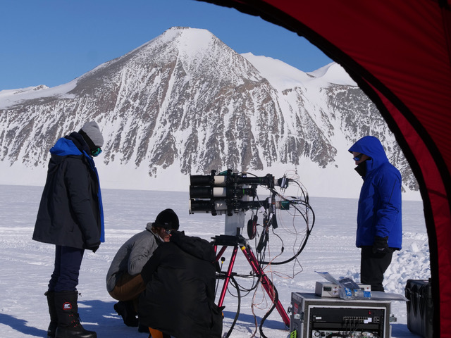 Four people wearing parkas work with photography equipment in Antarctica. A snow-covered mountain rises in the background. 