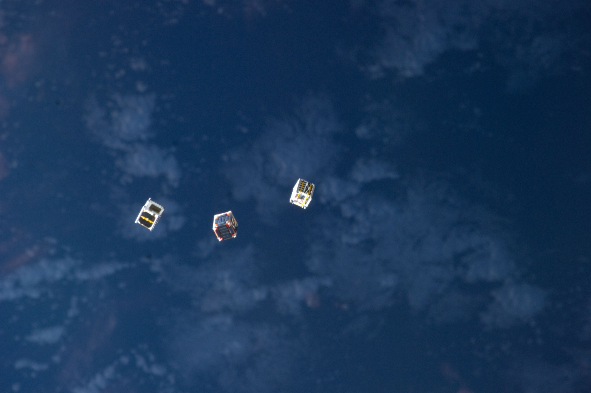 Three cubesats shown looking down from the international space station against a far off background of Earth's surface.