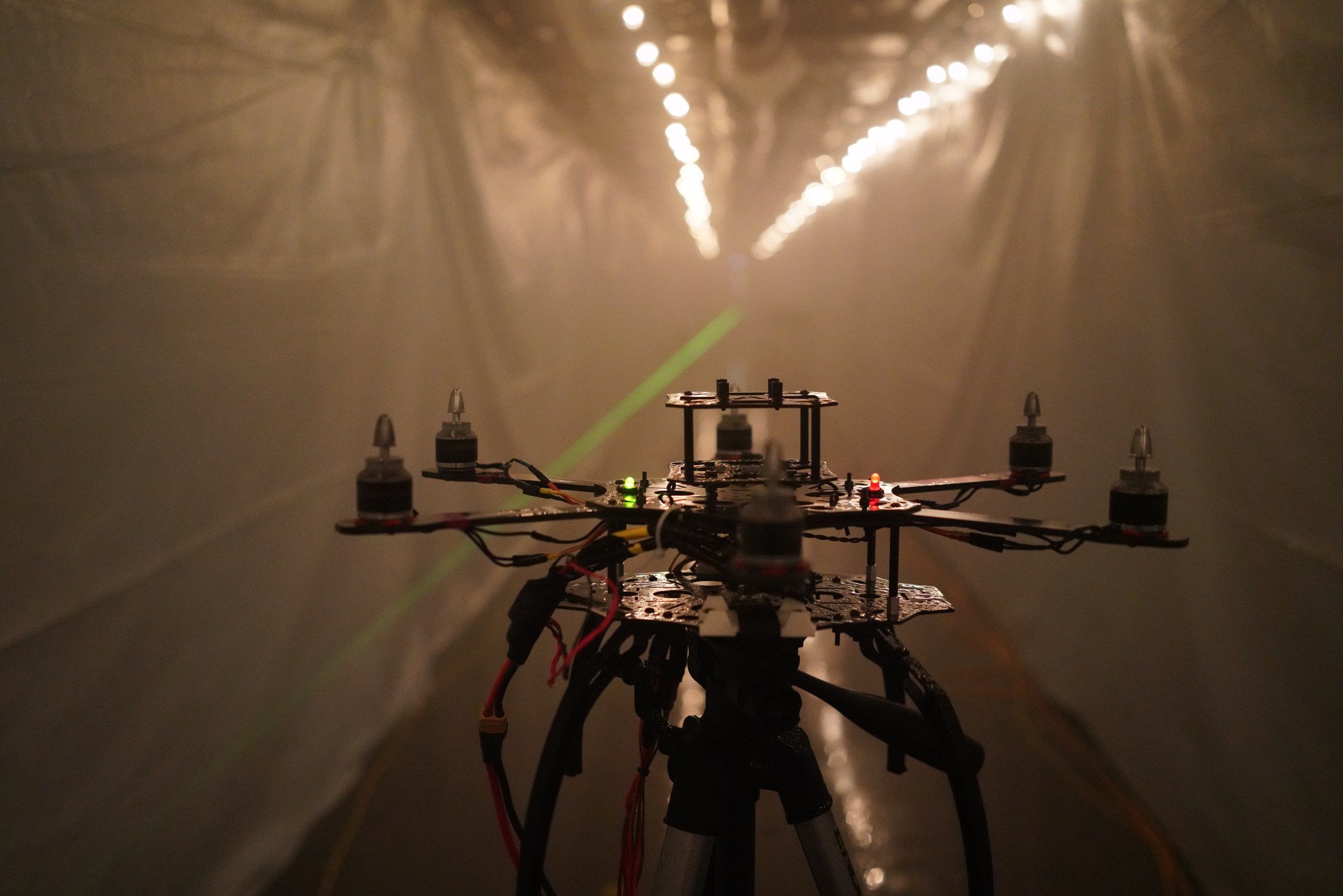 The frame of a drone sits at one end of a long passage filled with fog. Plastic sheeting covers the walls and two strings of lights line the ceiling.