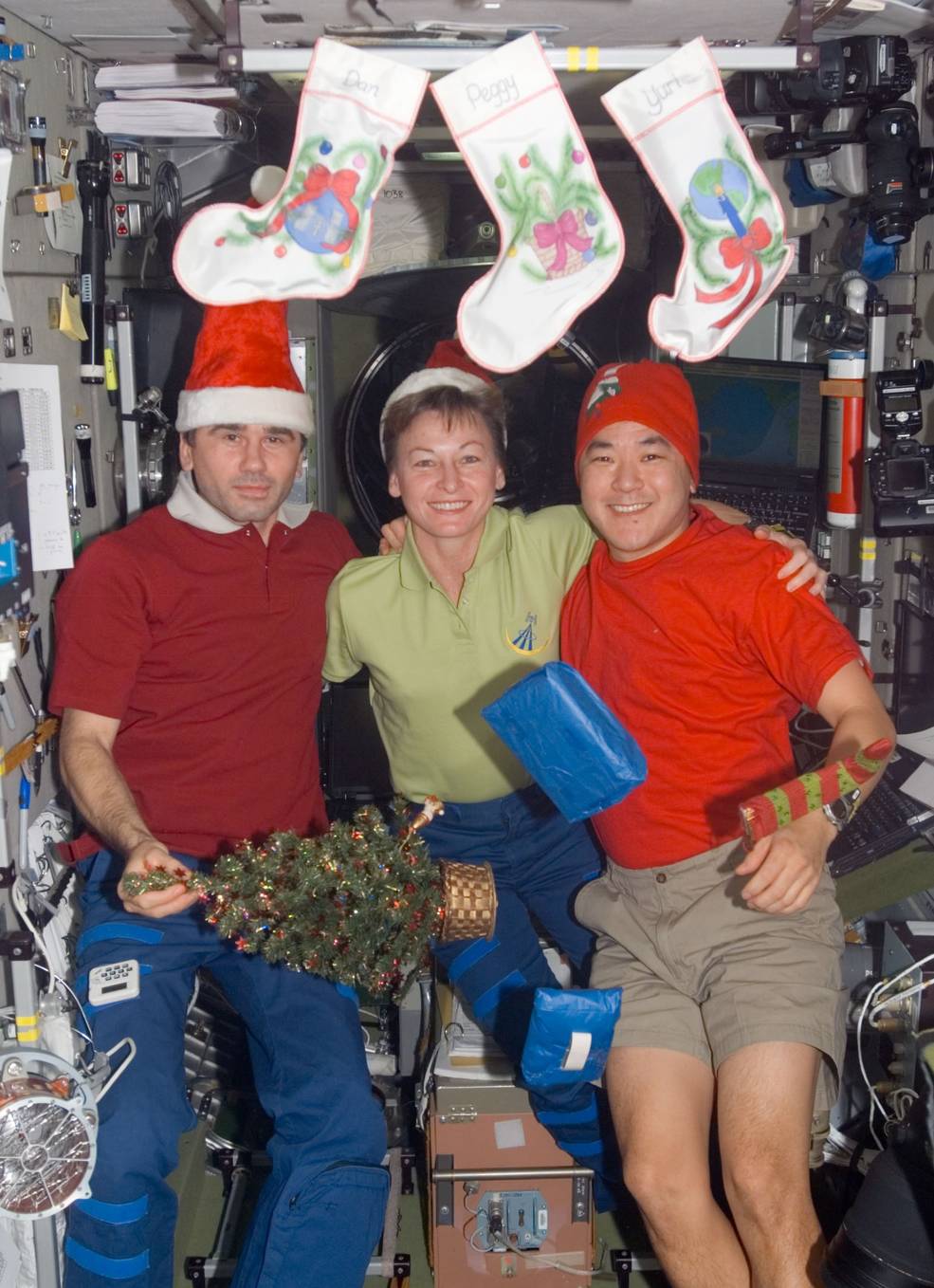 Two men and a woman wearing Santa hats pose in front of a set of Christmas stockings