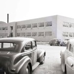 vintage black and white view of the administration buidling from the parking lot from between two 1940s automobiles.