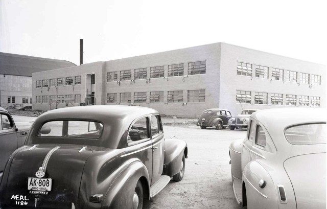 vintage black and white view of the administration buidling from the parking lot from between two 1940s automobiles.