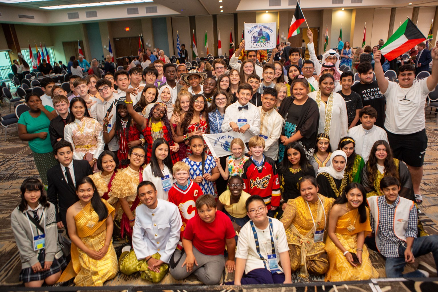 A photo of a large group of students, representing many different cultures, genders, and ages, posing and smiling in a large conference room. Many of the students wear traditional clothing from their cultures and hold national flags. The room has tan patterned carpet, and more national flags line the walls.