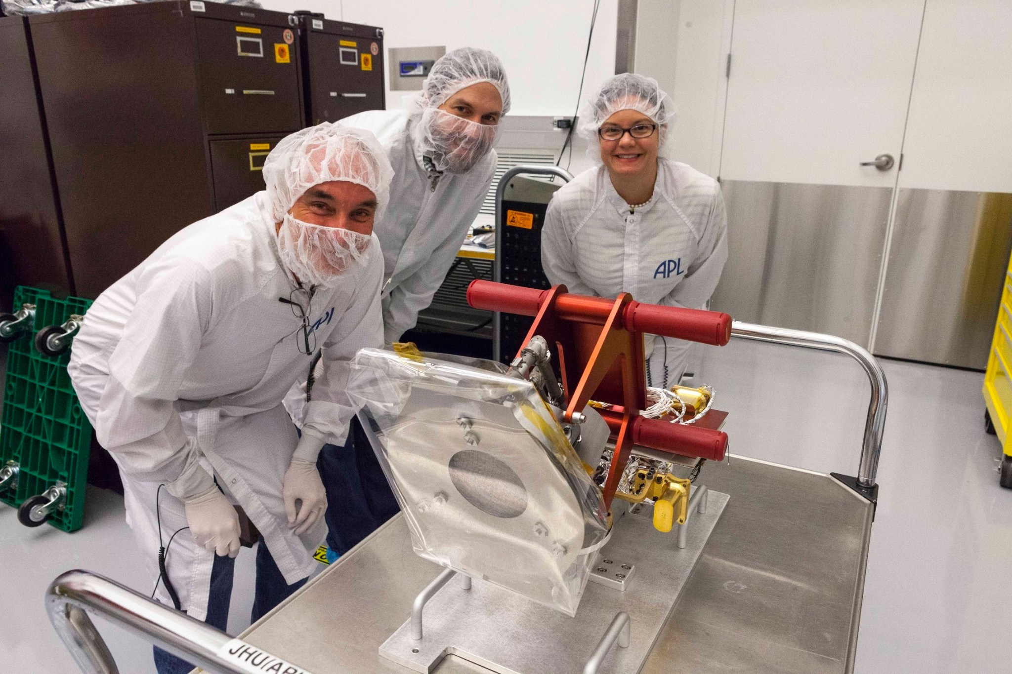 Three people in white scrubs and hairnets smile next to a scientific instrument. 