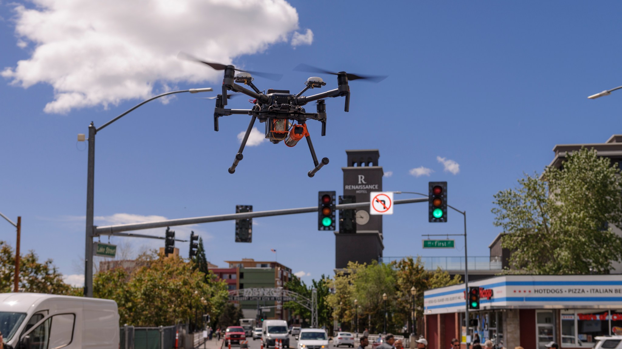 A black drone with four rotors flies above a city street, with traffic lights, buildings, and cars in the background.