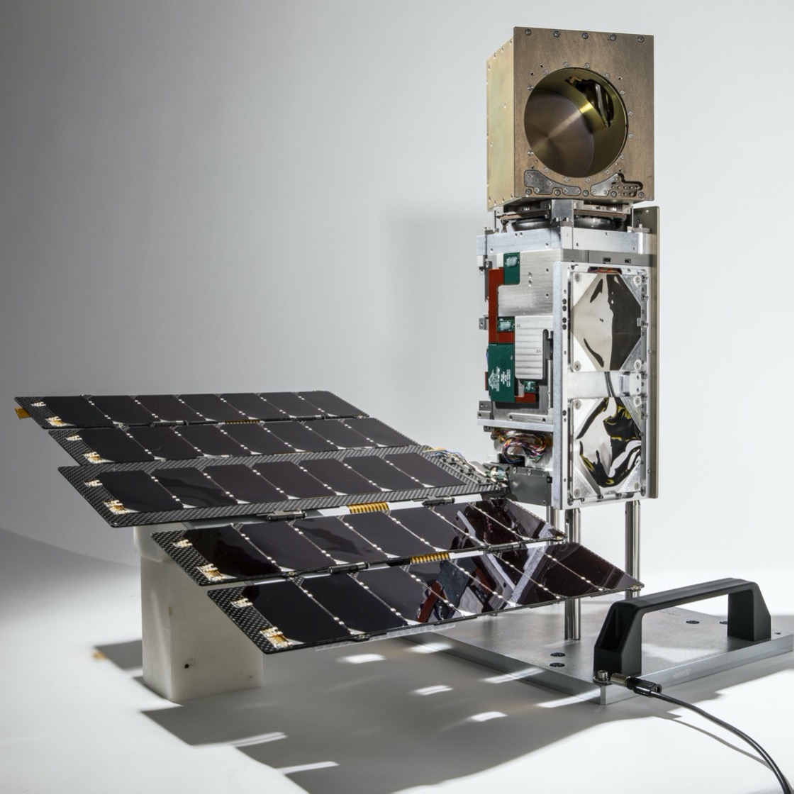 Photograph of the rectangular TROPICS CubeSat Pathfinder satellite standing on one end with its solar panel perpendicular and horizontal to its left at the base.
