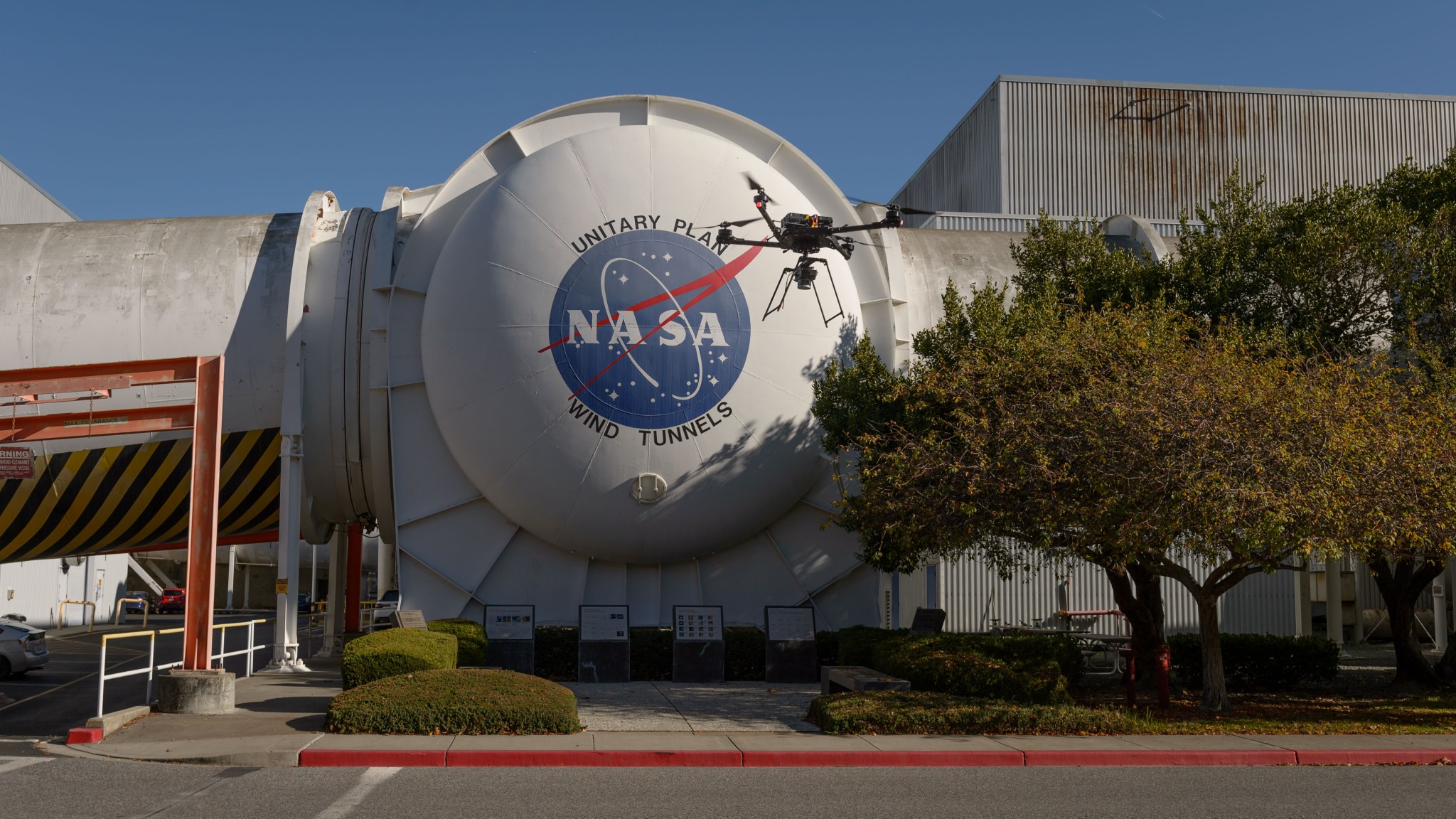 A quadcopter drone flies in front of the exterior of a wind tunnel bearing the NASA logo