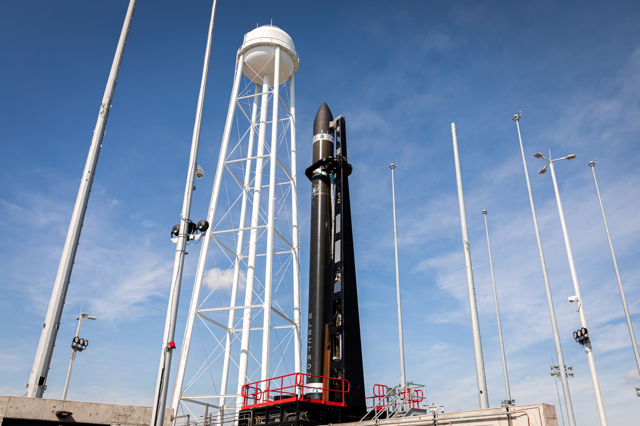 All black rocket on pad with the words "Electron" in white vertically down the rocket with blue skies and wispy clouds in the background.