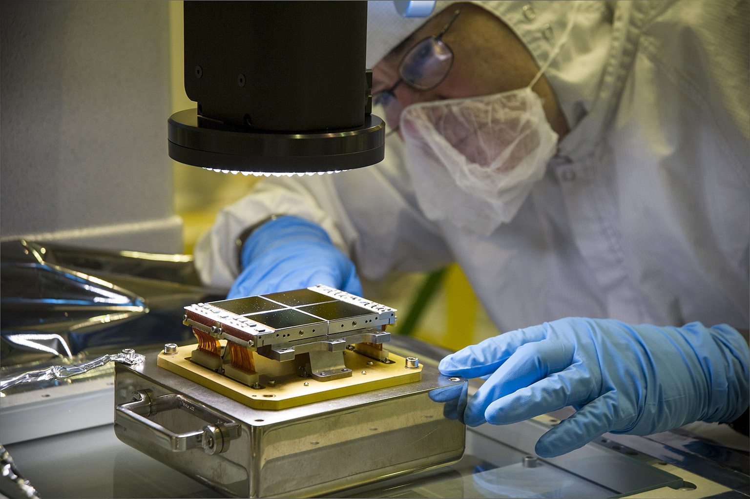 A technician in a cleanroom suit inspects parts to an instrument through a 3D microscope.