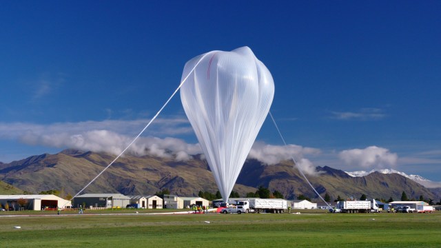 A scientific balloon, clear and in the shape of an upside down teardrop, is partially inflated before launch. A mountain with light clouds is in the background.