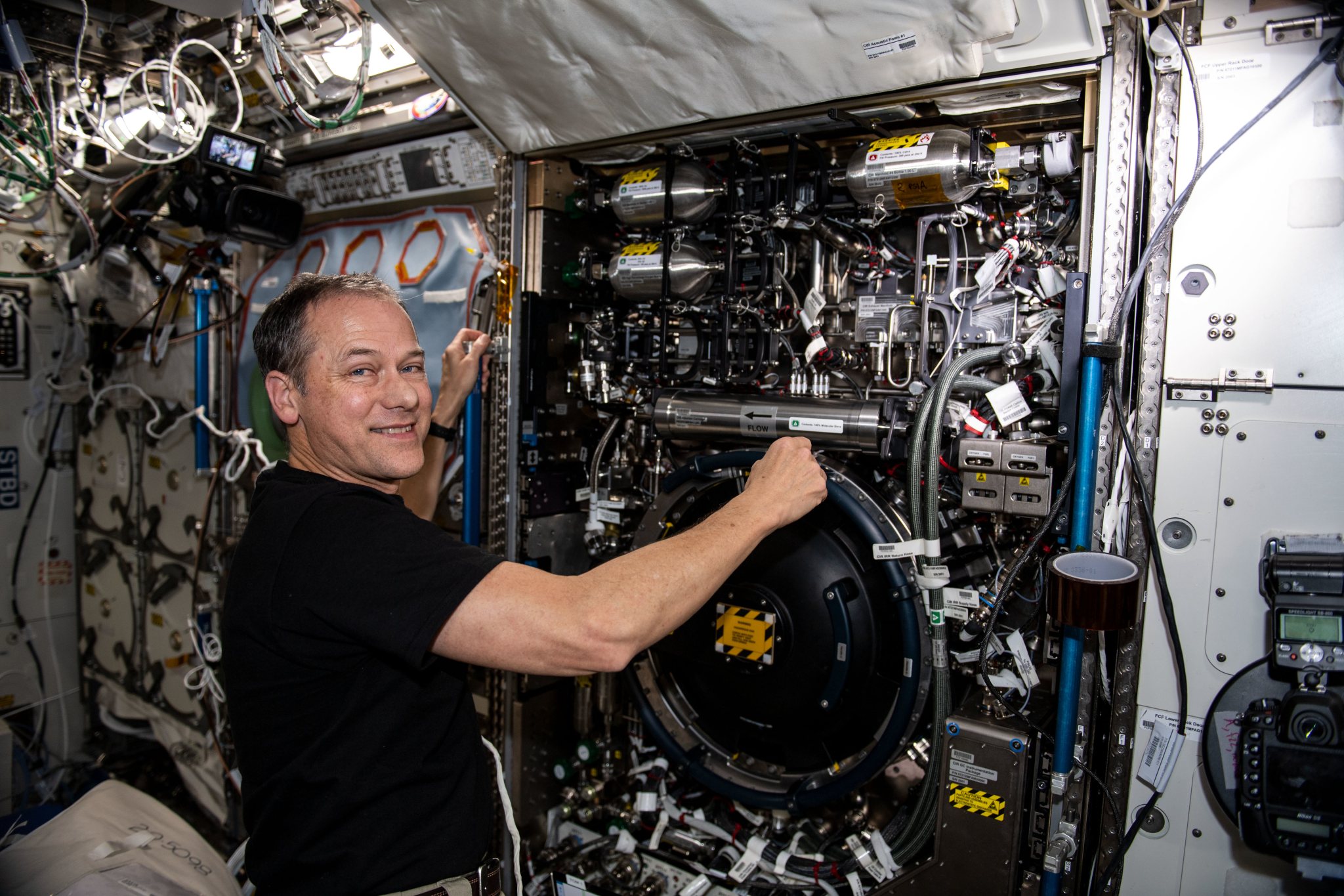 Alt text: Astronaut looks at camera as he works near a combustion experiment rack.