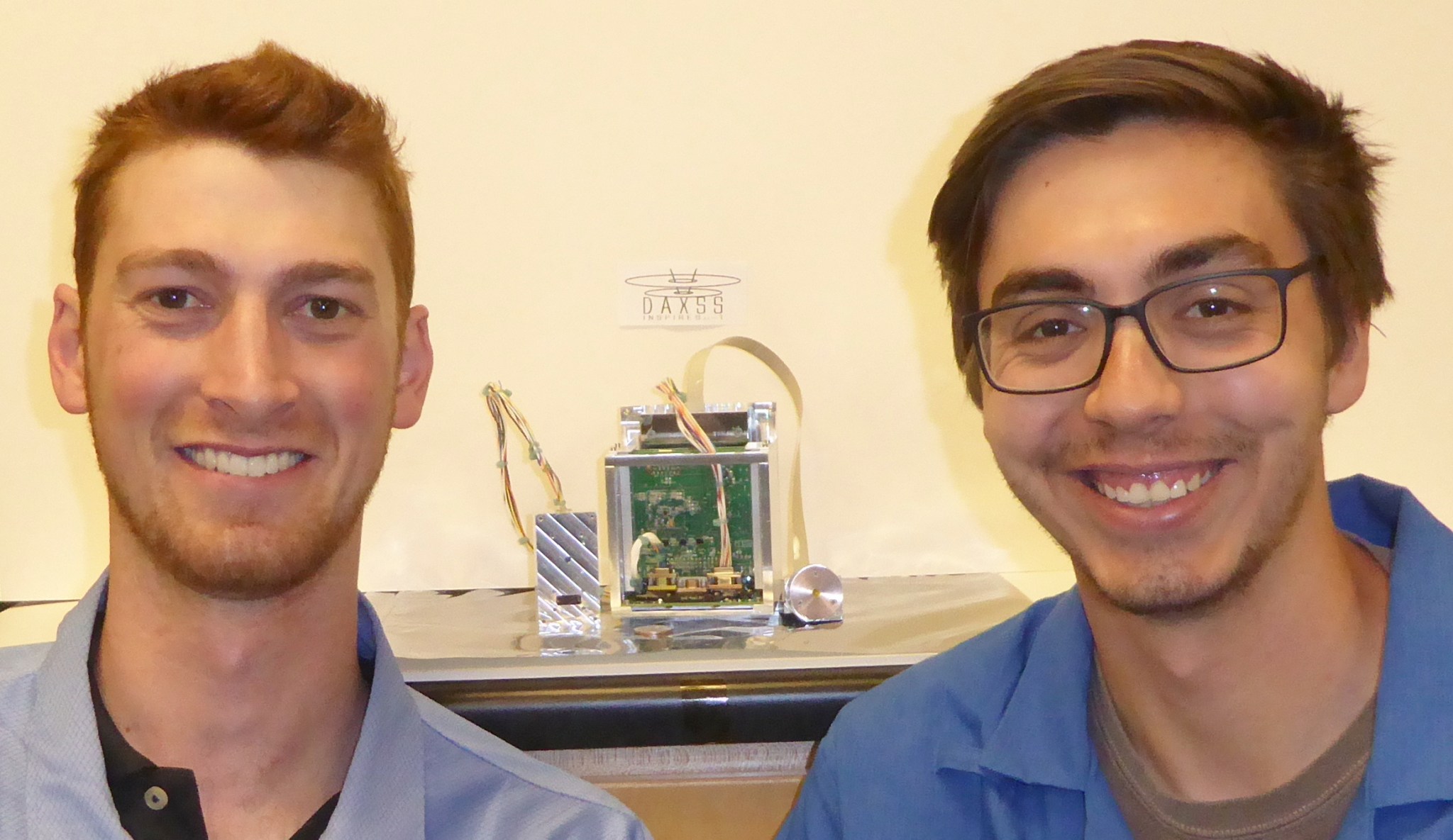 Two young men smiling with a piece of machinery in the background.