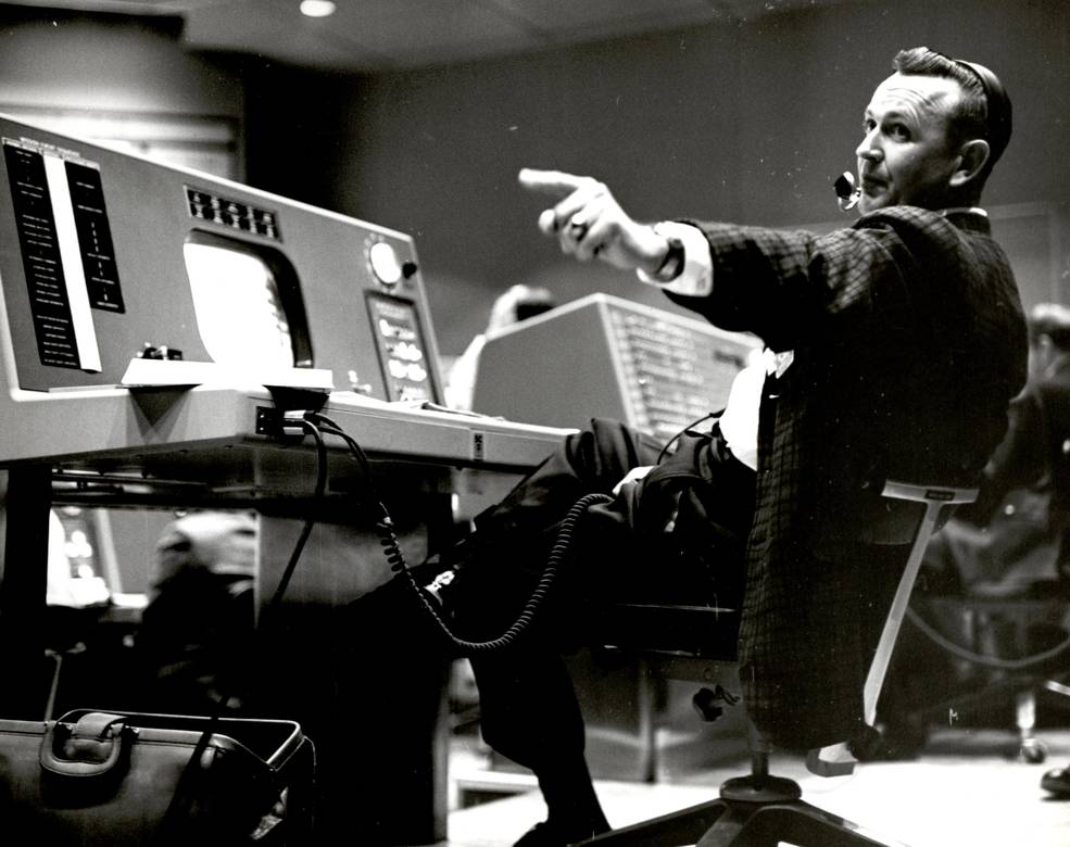 Christopher Kraft, flight director during Project Mercury, leans back in his chair and points with his left hand at his console inside the Flight Control area at Mercury Mission Control.