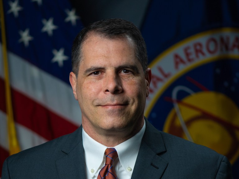 Joe Schuyler sits for a portrait wearing a dark suit and orange striped tie; Both the United States of America and NASA flags are visible behind him