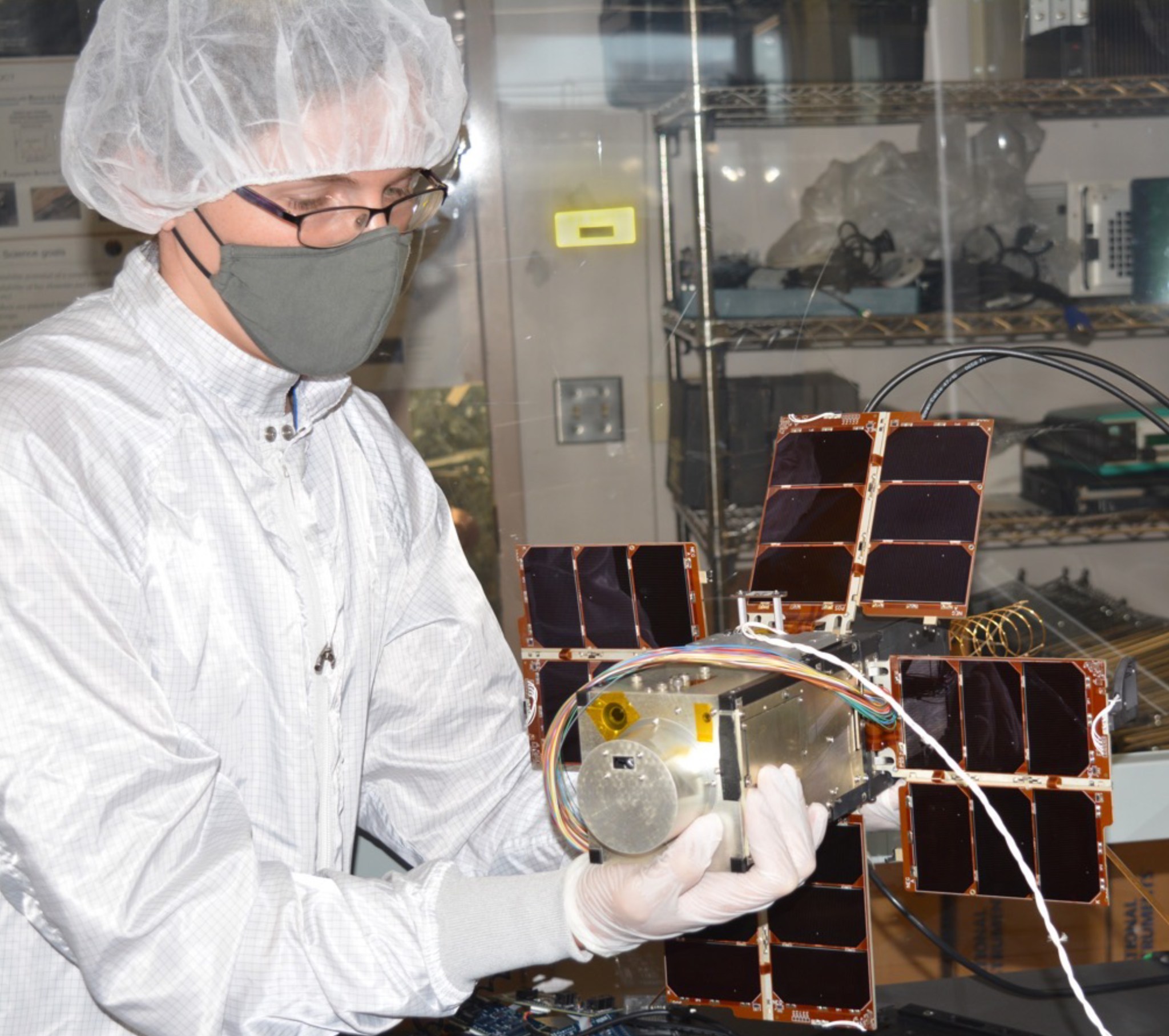 Person in white clean room attire holding a small, cube-like satellite. The satellite is silver and gold, with a squat cylinder protruding from one end. 