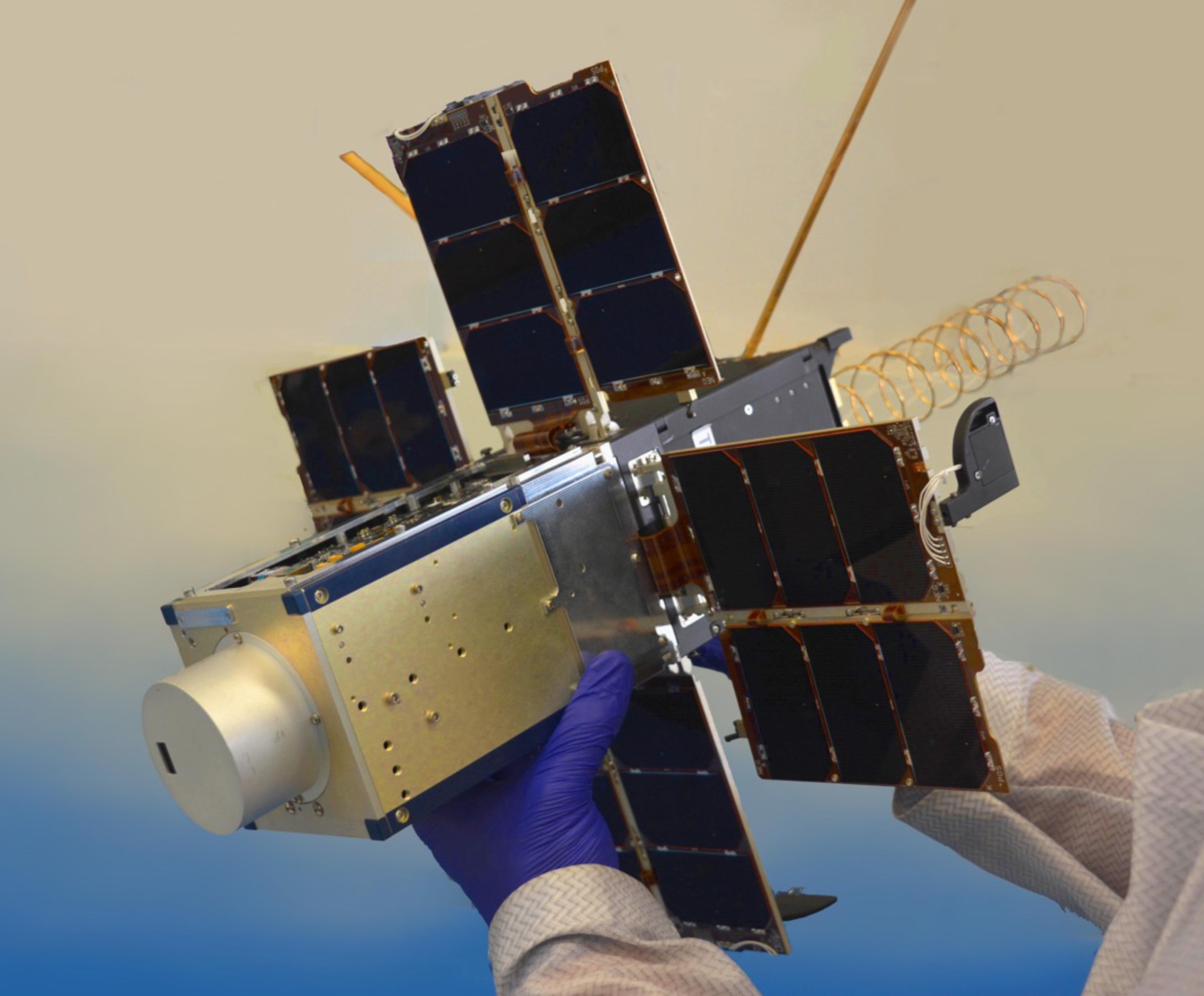 Blue gloved holds up NACHOS, a small, cube-like satellite with four solar panels. The CubeSat is silver and gold with a squat cylinder protruding from one end of the satellite.