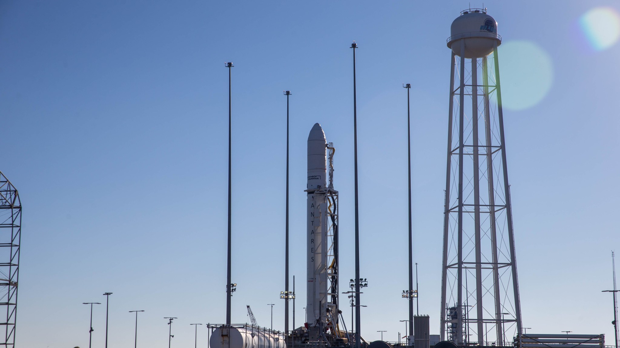 A large, white rocket stands on its launch pad. To the right of the rocket, a water tower with long, spindly white legs has a white dome on top, with a dark logo that’s not fully visible against a bright blue sky.