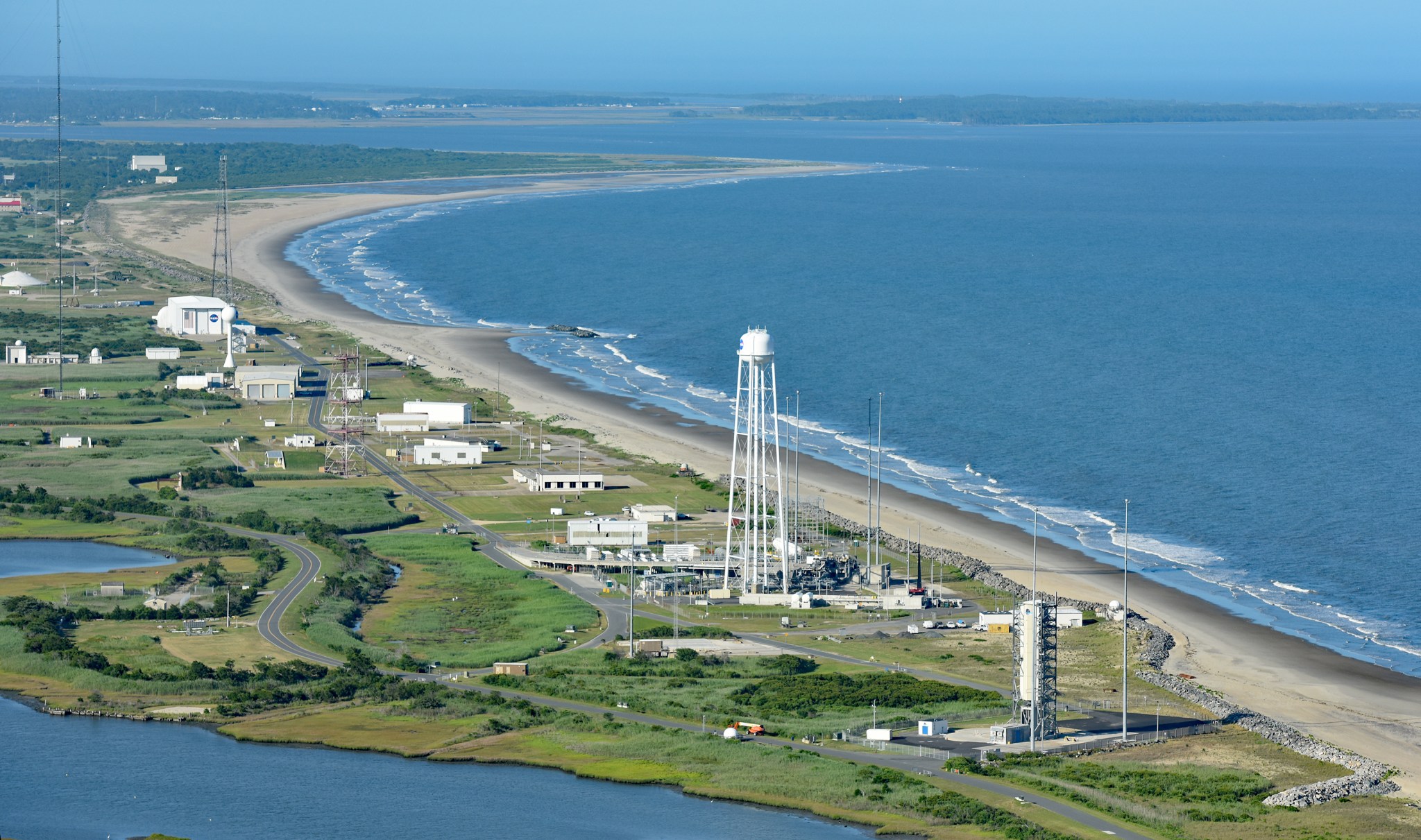 Aerial view of the coastal launch range of Wallops Flight Facility, showing a blue Atlantic Ocean on the right; white buildings along a tan coastline back up to a green, marshy landscape.