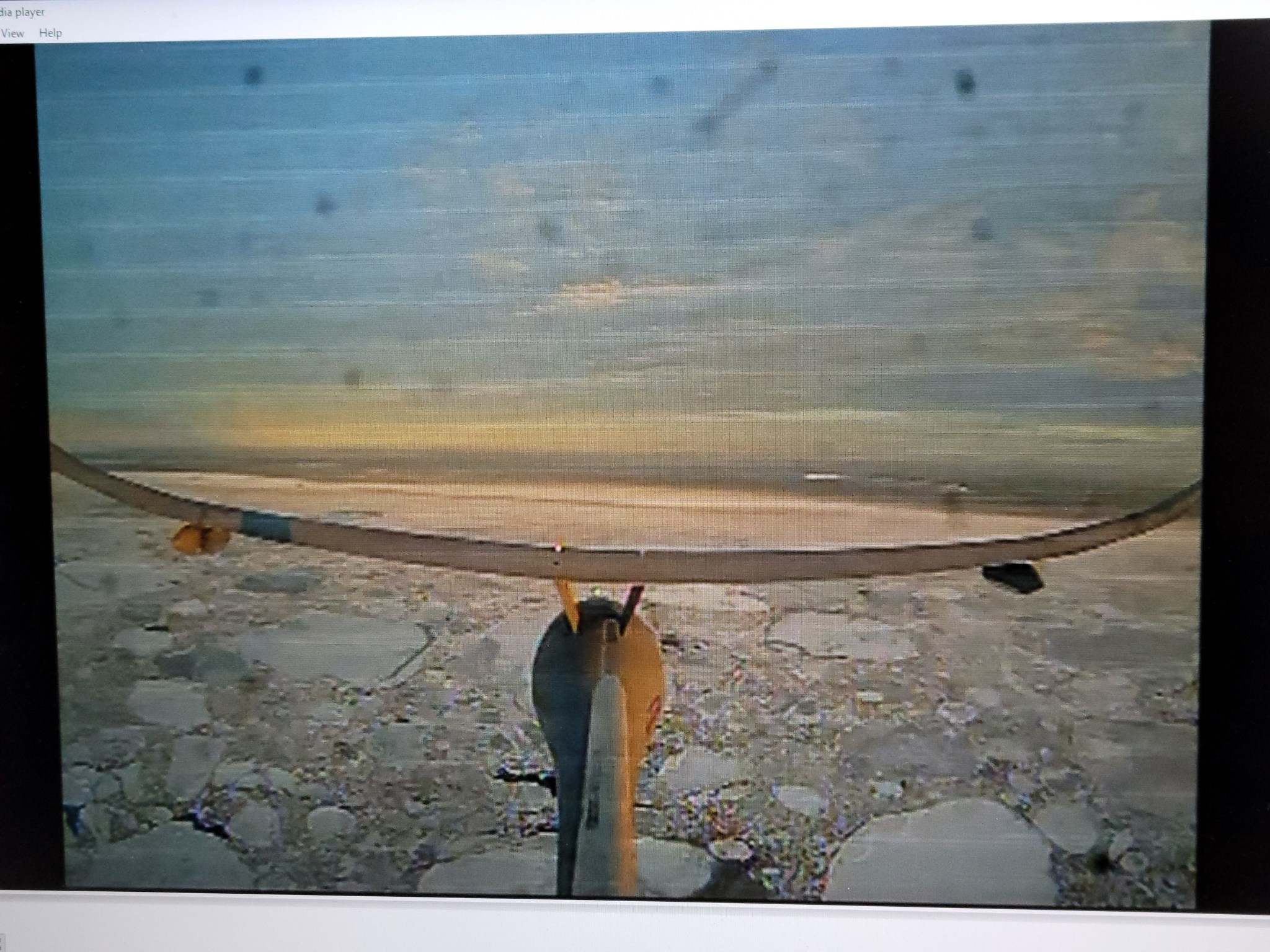 A drone tail camera shows Arctic sea ice. The drone's body and wings are in the foreground with chunks of white ice filling the water.