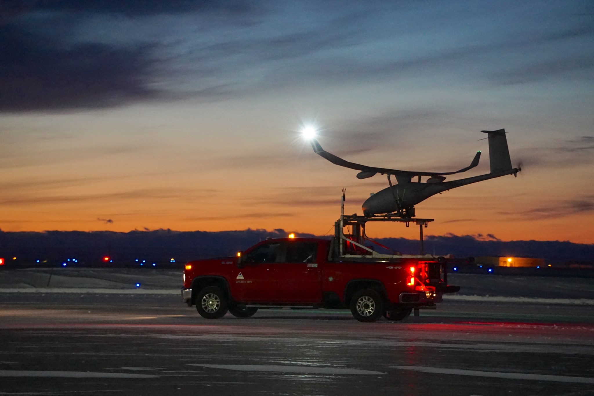 Fixed-wing drone prepares for takeoff in Deadhorse, Alaska. The drone looks like a tiny airplane mounted on the back of a red pickup truck. The sunset in the background makes the sky orange and light blue.