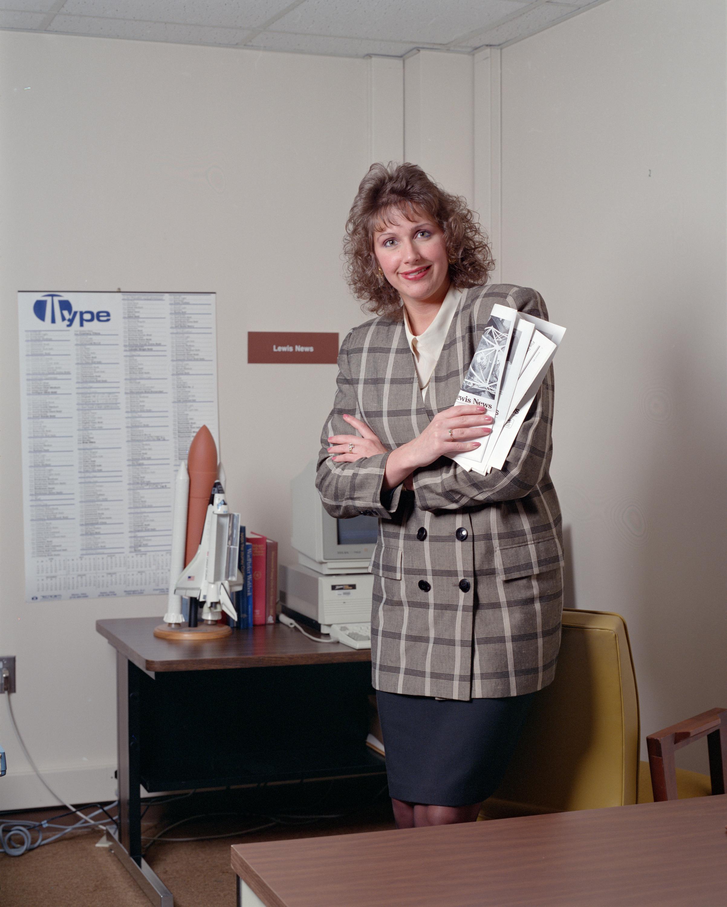 Woman looking at the camera stands in front of a desk topped with an old computer and space shuttle model.