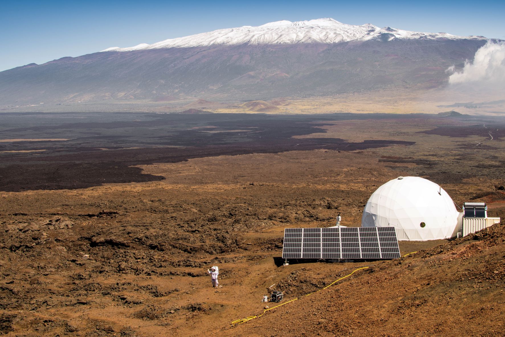 A view of the the NASA-funded Hawaii Space Exploration Analog and Simulation, or HI-SEAS, facility. In the foreground is a crew member simulating a walk on a planetary surface. In the background is Mauna Loa.