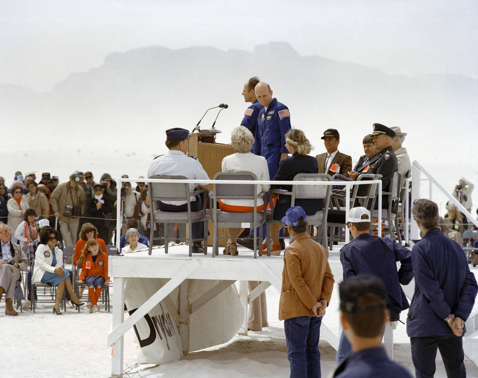 Astronauts Jack Lousma and C. Gordon Fullerton stand at a podium on a stage set up at White Sands Missile Range to address a crowd that has come to greet them on their return from STS-3