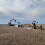 Construction equipment in a dried marsh area. There are three vehicles lined up in a row cutting a line through the marsh. Personnel are holding long, black cable lines that are being buried into the ground.