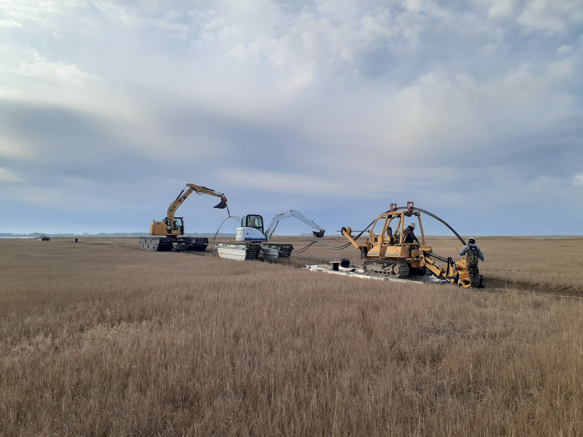 Construction equipment in a dried marsh area. There are three vehicles lined up in a row cutting a line through the marsh. Personnel are holding long, black cable lines that are being buried into the ground.