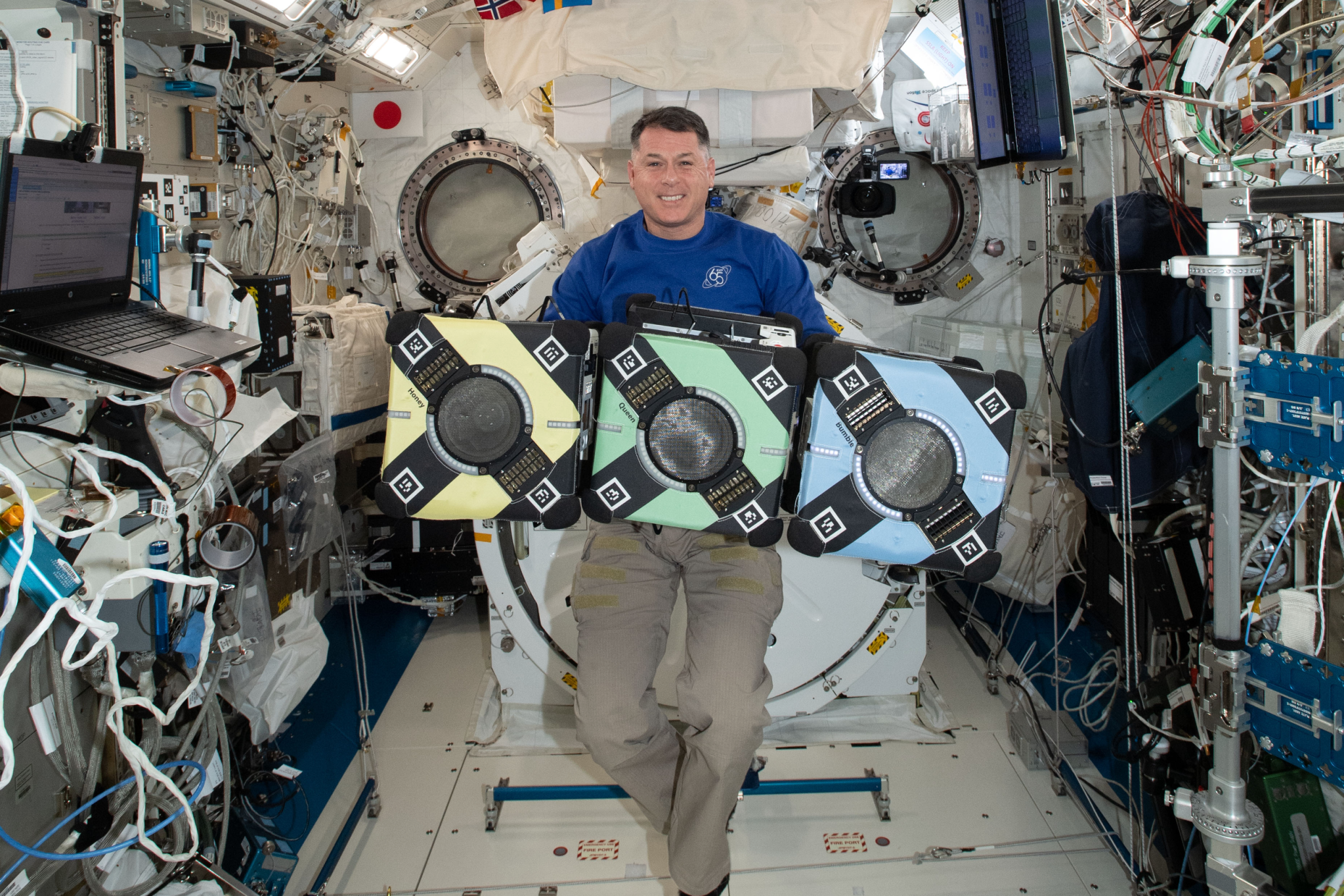 Astronaut in a blue polo floating behind three cube-shaped floating robots, colored yellow, green, and blue from left to right. All are in a hallway of the International Space Station.