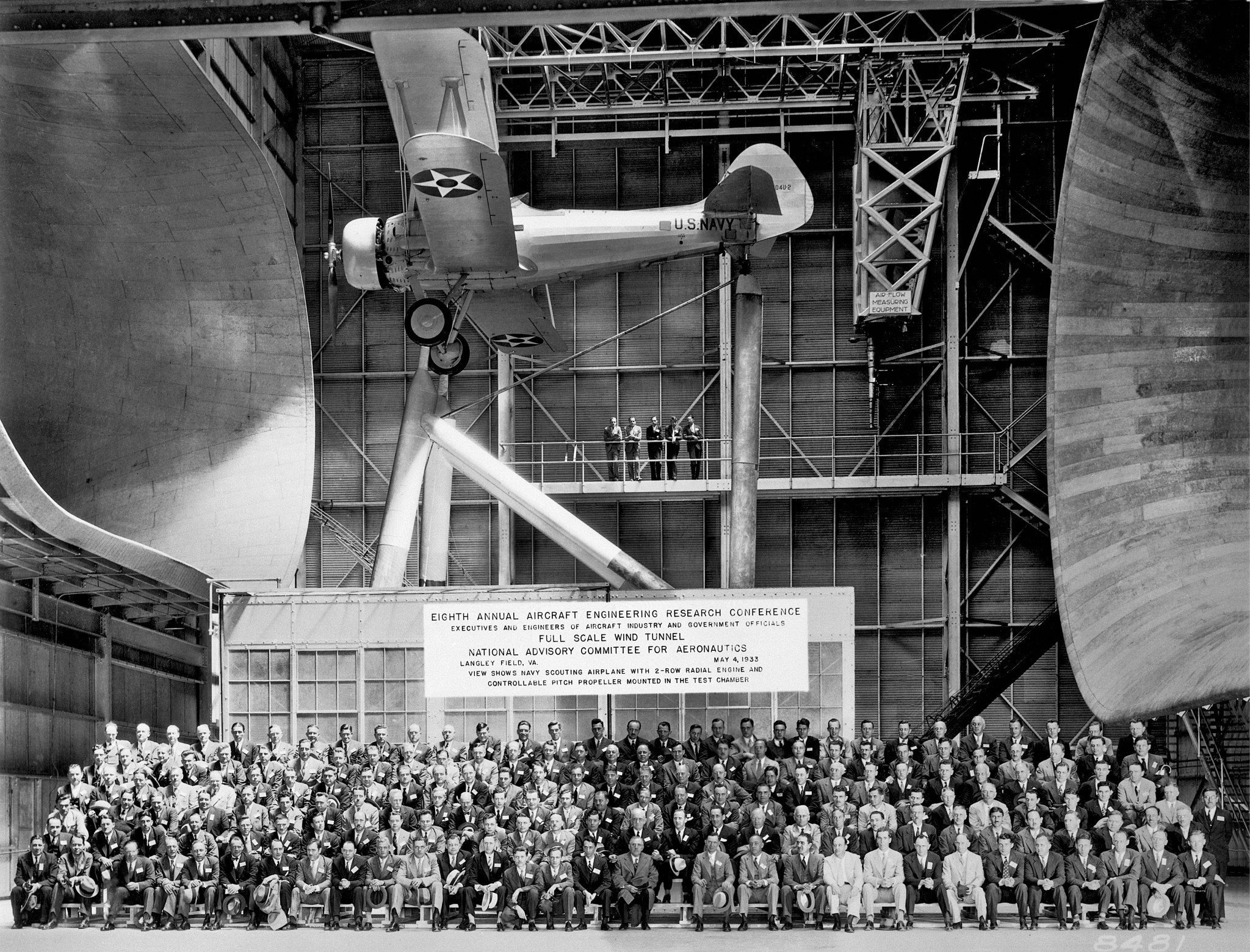 This is a photo of a large group of people, attendees at the 8th Annual Aircraft Engineering Research Conference, sitting in bleachers in the Full-Scale Tunnel. A U.S. Navy scouting aircraft hangs from the ceiling above the attendees.