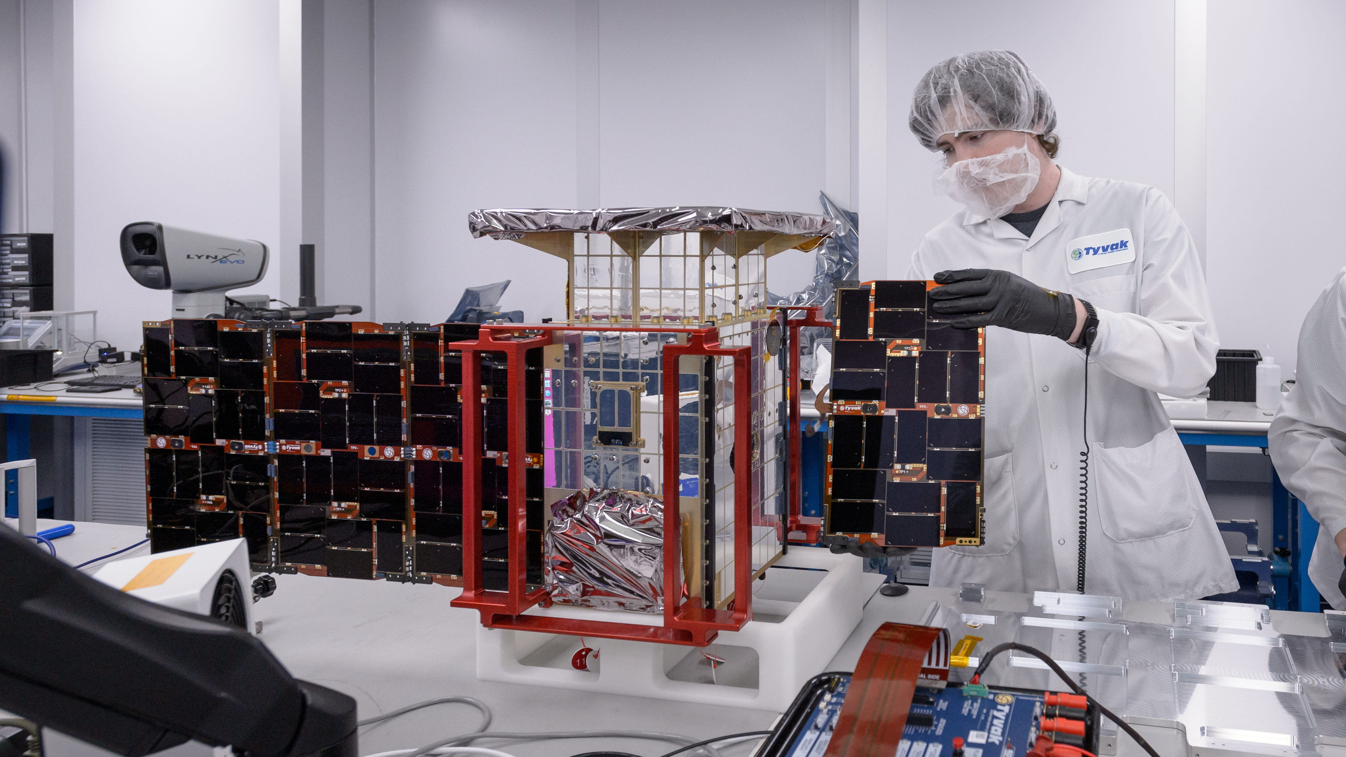 An engineer stands behind a small satellite with one solar panel unfurled and the other half being installed.