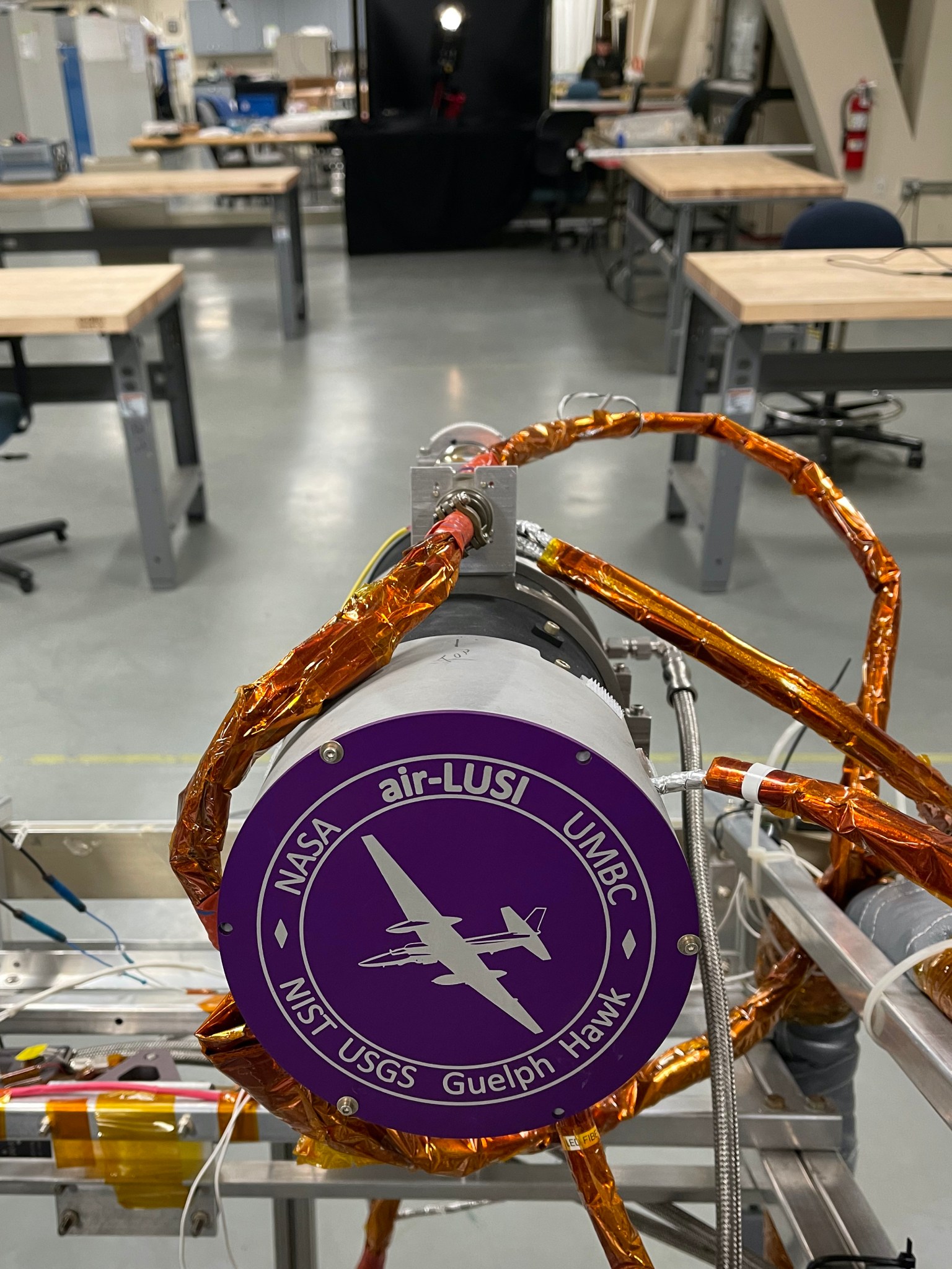 The cylindrical air-LUSI telescope is positioned to measure a simulated Moon at the far end of a laboratory. The instrument is a cylinder with a purple label on the front that says NASA air-LUSI, UMBC, NIST, USGS, Guelph Hawk. Bronze wires extend from the instrument.
