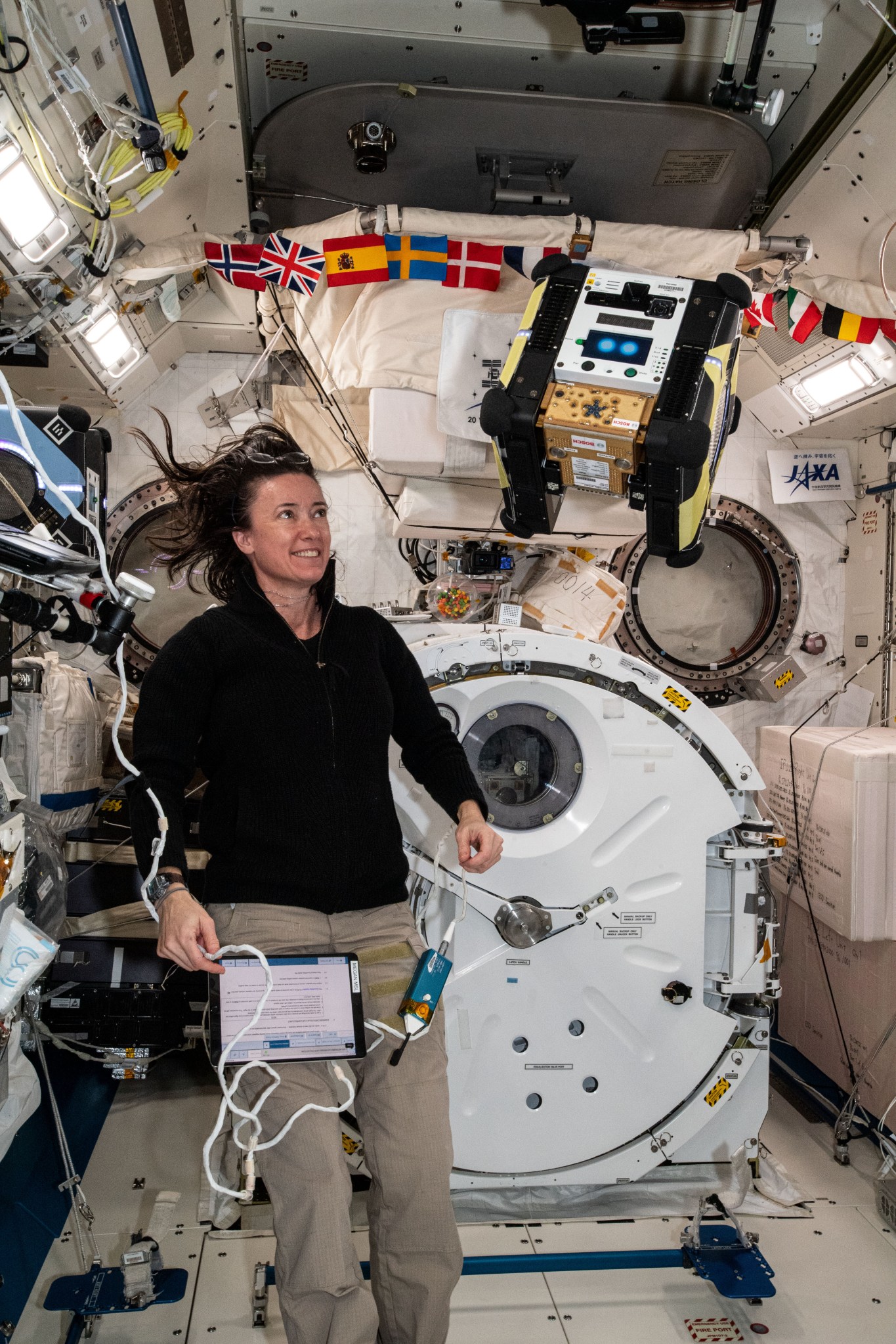 Astronaut floating in a corner of the International Space Station with a cube-shaped floating robot with yellow and black panelling. There is a circular door behind them.