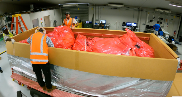 Two man in bright orange vests stand around a large container. Inside is the deflated super pressure balloon covered in bright orange plastic.