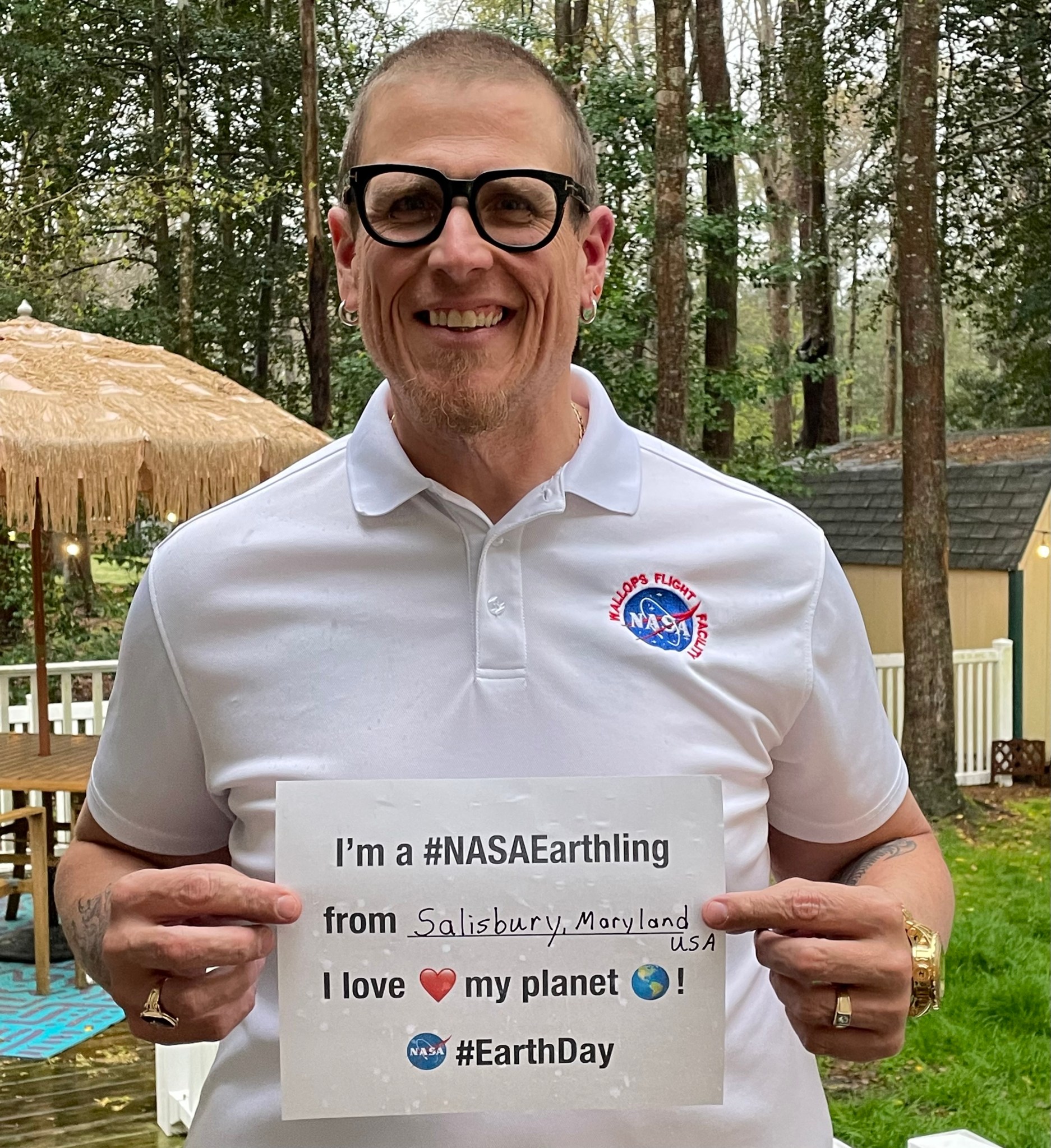 A man with buzzed hair, glasses, and earrings holds a paper sign outdoors. The sign says, “I’m a #NASAEarthling from Salisbury, Maryland, USA. I love my planet! NASA logo, #EarthDay” with a heart emoji, an Earth emoji, and the NASA meatball logo.