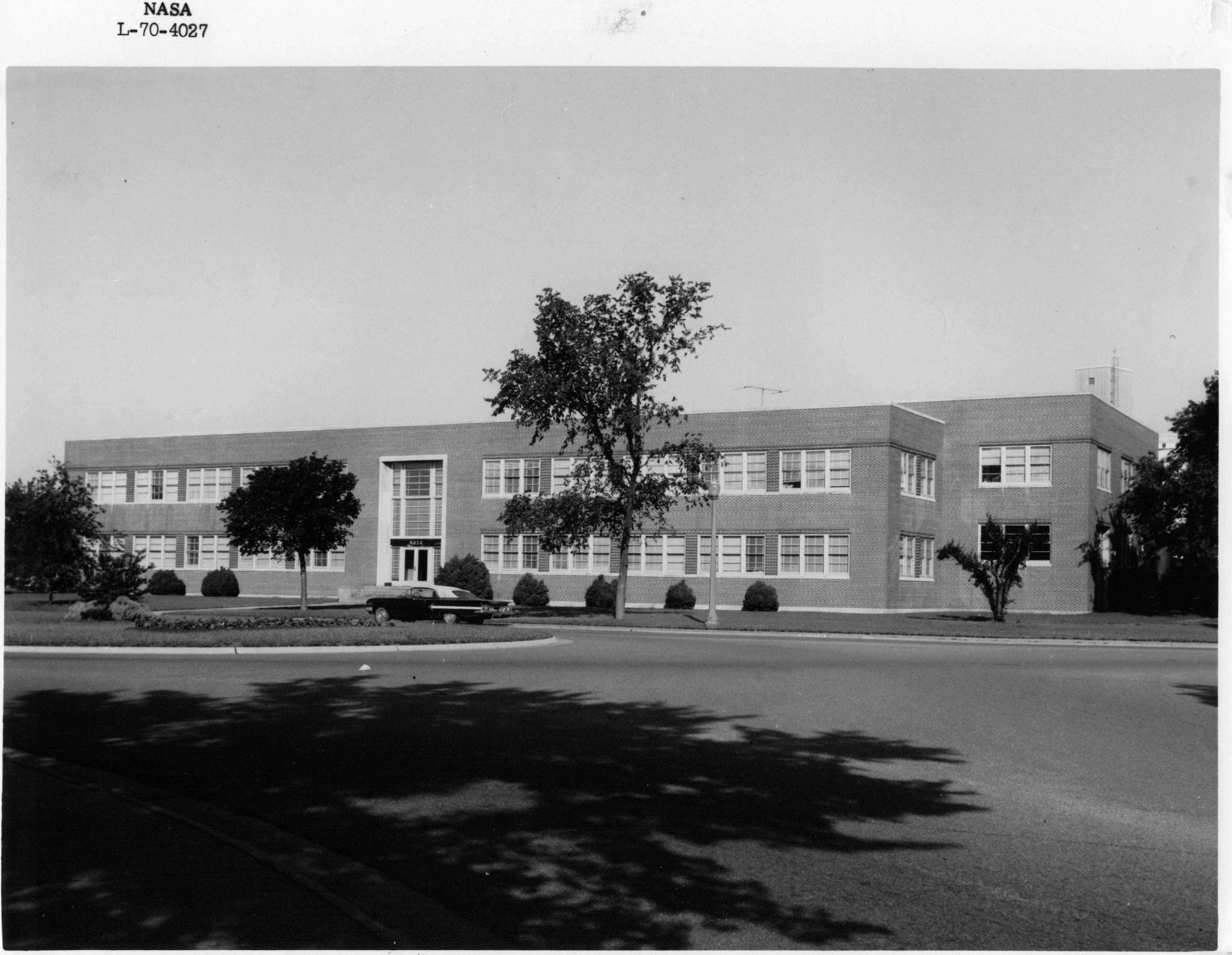This is a photo of the exterior of Building 1229 taken in 1970. Trees and bushes line the street and a retro car is parked in front of the building.