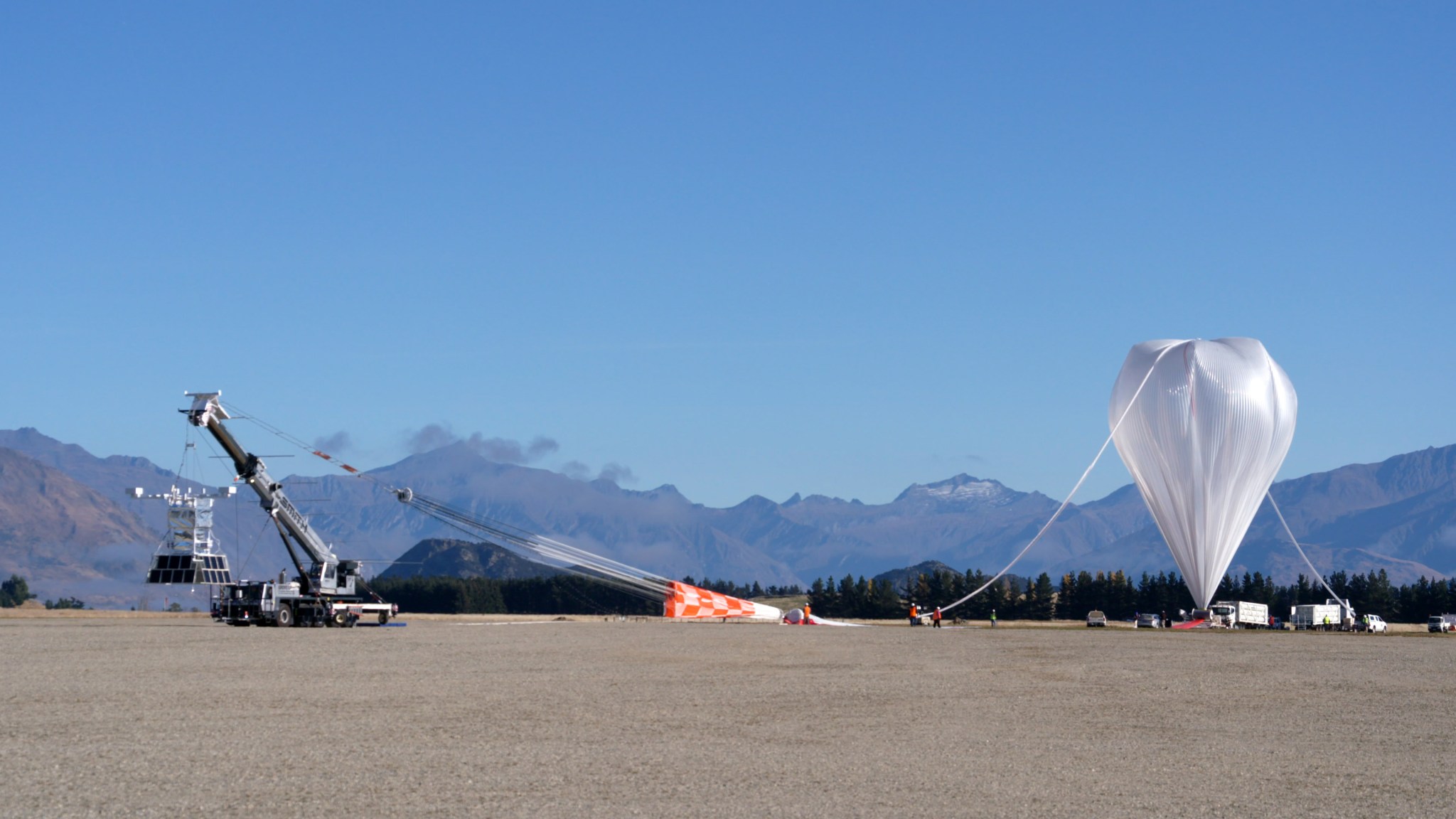 A crane is leading a scientific balloon before launch. The balloon is to the right, and appears an a plastic, upside down teardrop. A tube attached to the top of the balloon leads down to the ground where a number of personnel are holding it. To the left, a crane holding a large payload structure with many solar panels is attached to the end of the balloon.