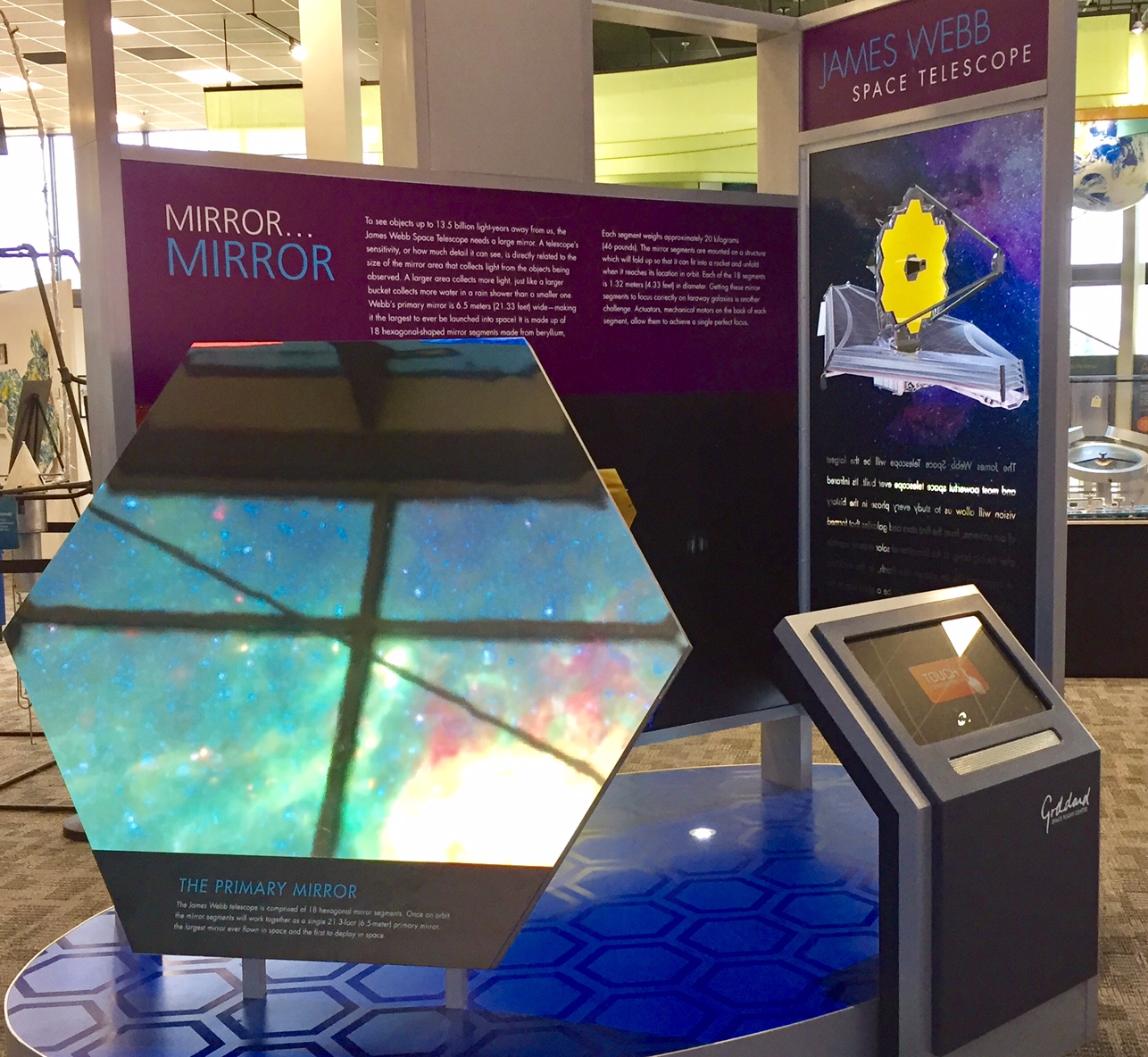 An exhibit about the James Webb Space Telescope. In the background are two boards with information about the telescope. One reads Mirror…Mirror. The other says James Webb Space Telescope. In the foreground, there’s an example of the gold hexagon mirror. 