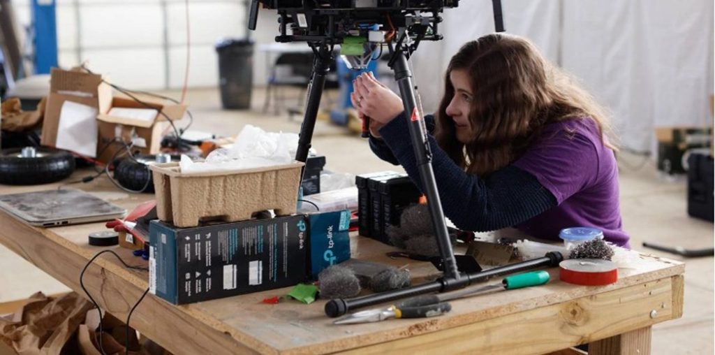 A researcher works to assemble a small spacecraft technology payload perched on a wooden tabletop in a ground-based technology facility.